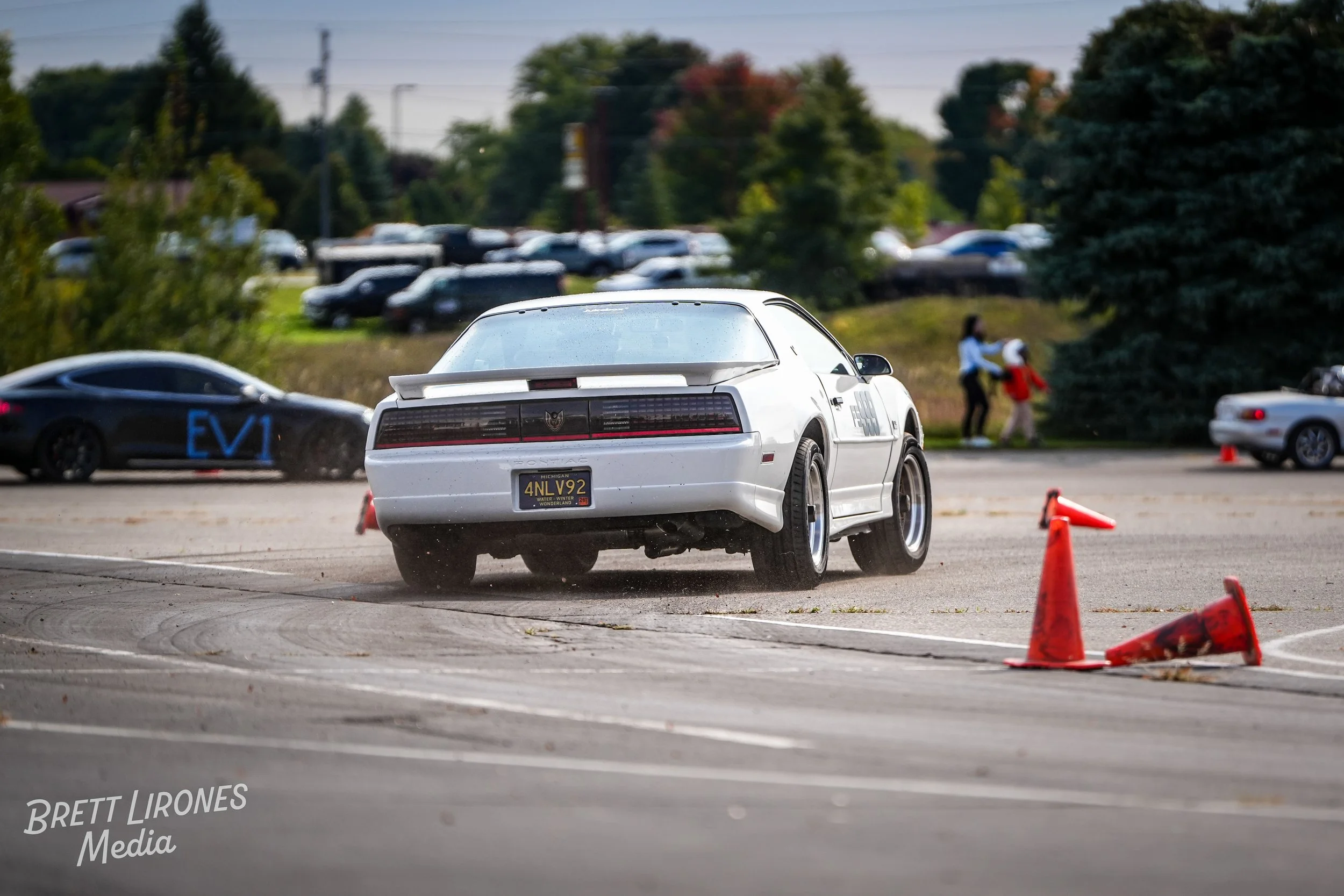 White classic car taking a sharp turn on a paved area, with orange cones knocked over, in a parking lot with trees and people in the background.