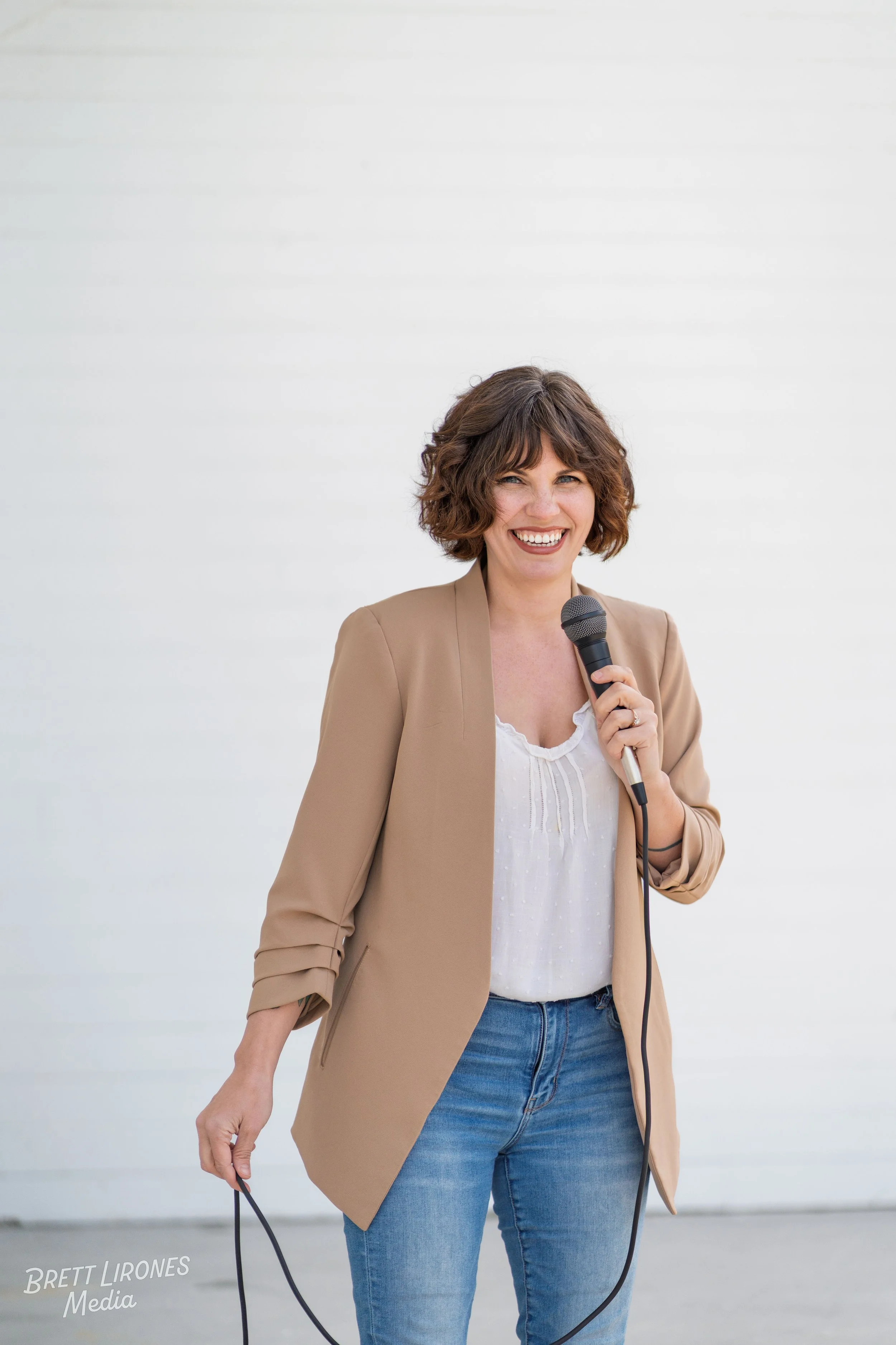 A woman smiling and holding a microphone, wearing a beige blazer and blue jeans, standing against a plain white background.