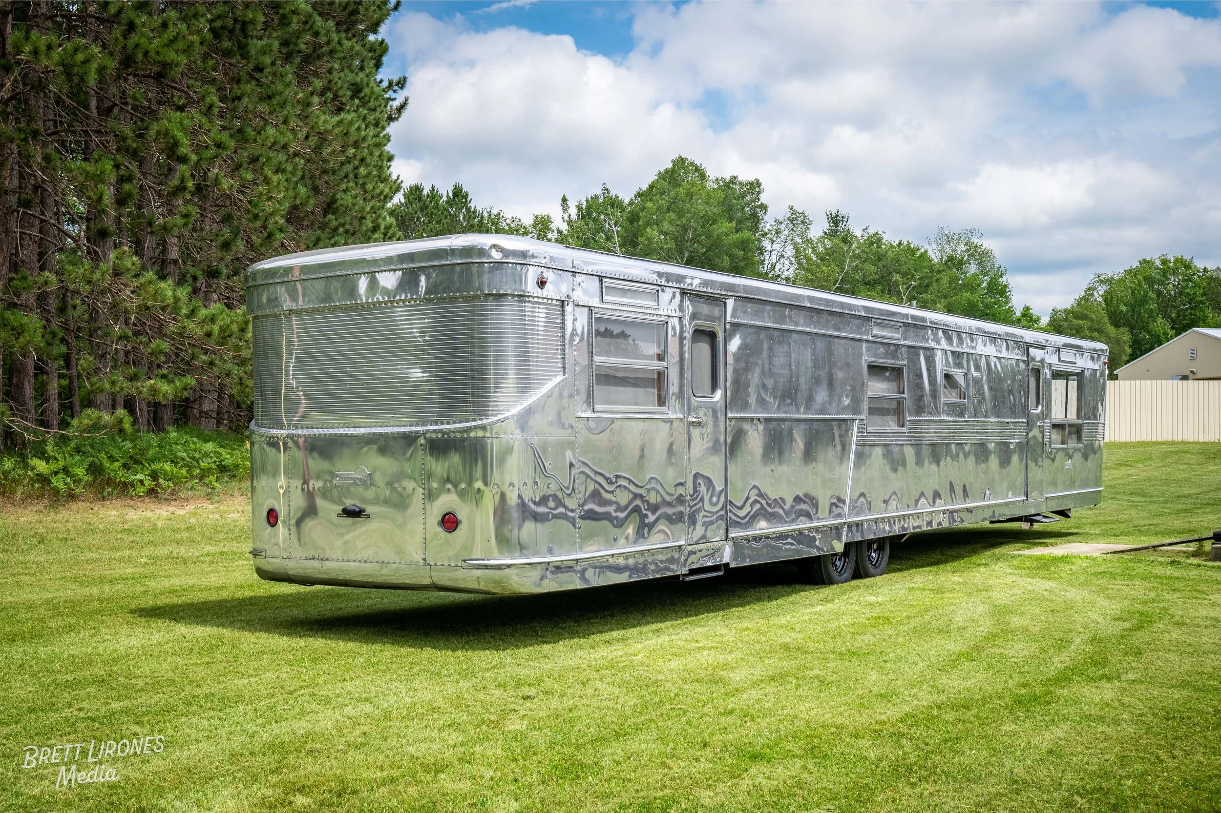 A shiny, vintage silver travel trailer parked on a grassy field with trees and a house in the background.