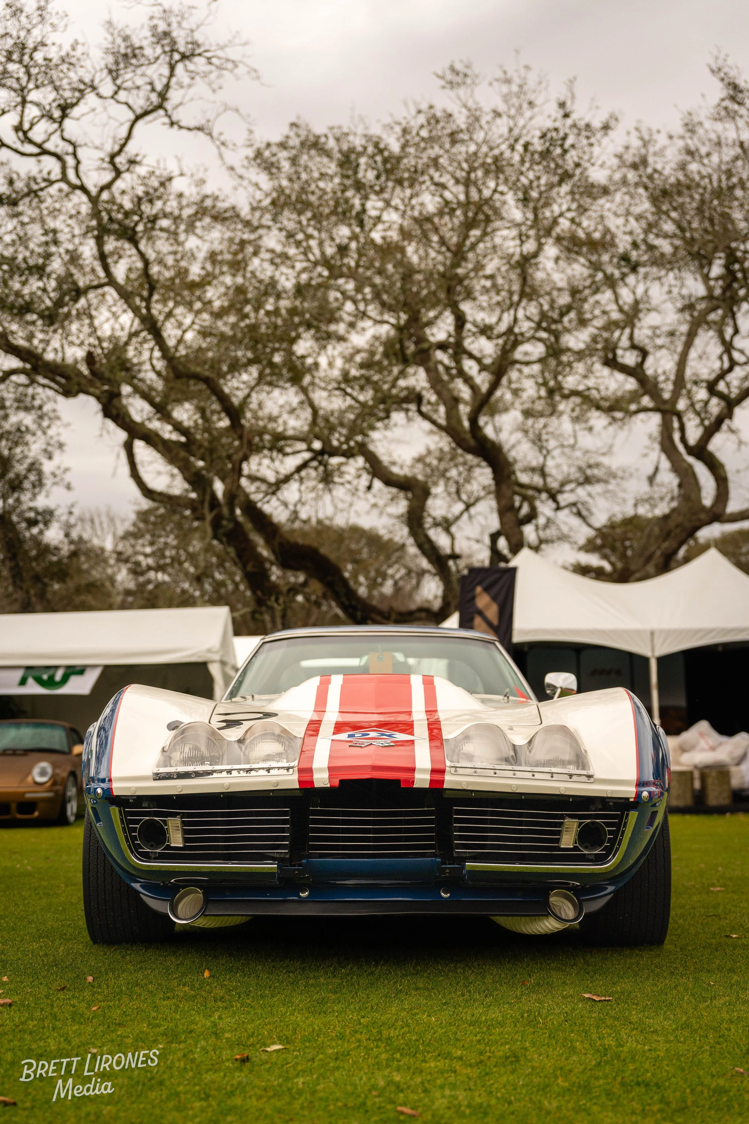 A vintage race car with a white body, blue and red stripes, and racing decals, parked on a grassy area at an outdoor event with tents and trees in the background.