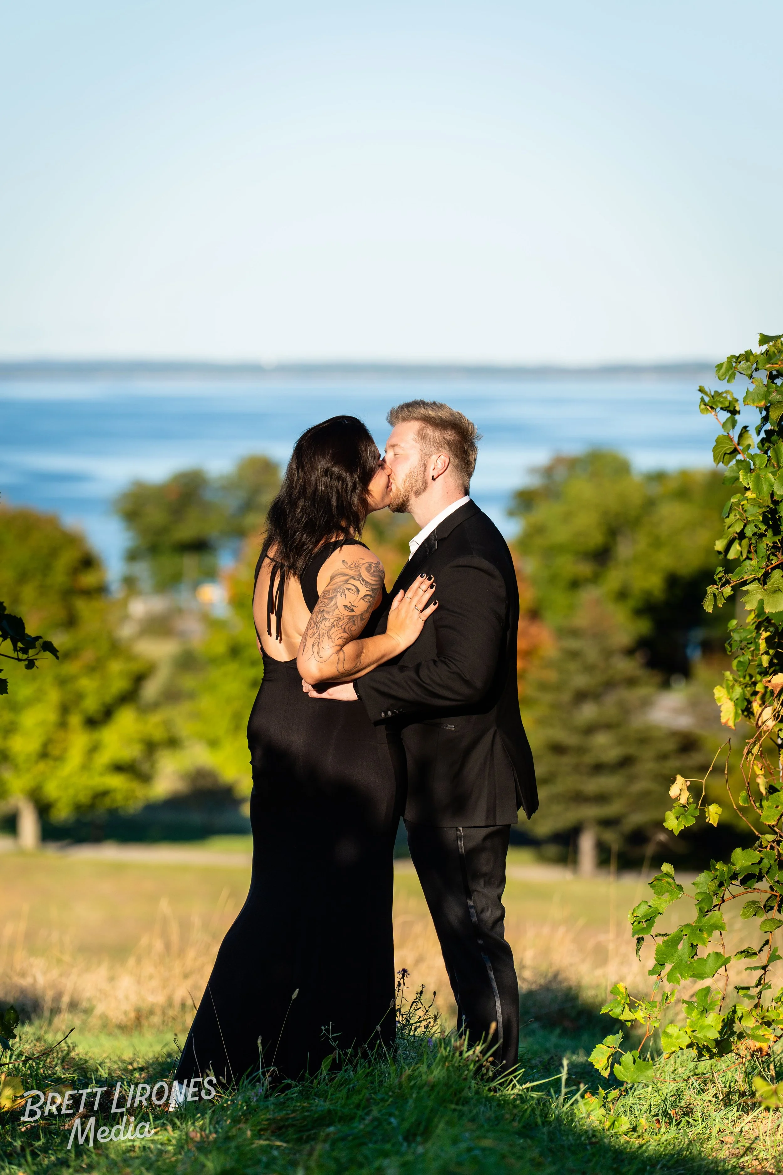 A couple dressed in formal attire sharing a kiss outdoors near a lake with trees in the background.