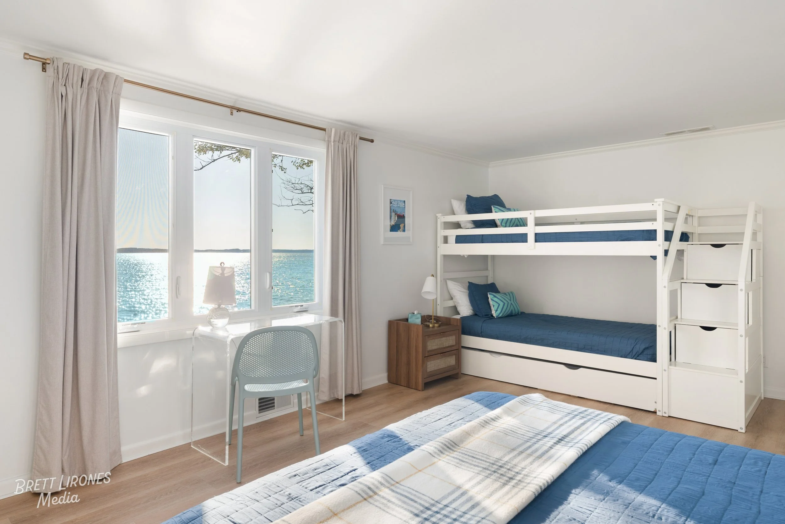 A bedroom with a bunk bed, blue bedding, a large window showing a view of the water, and a desk with a chair and a small table lamp.