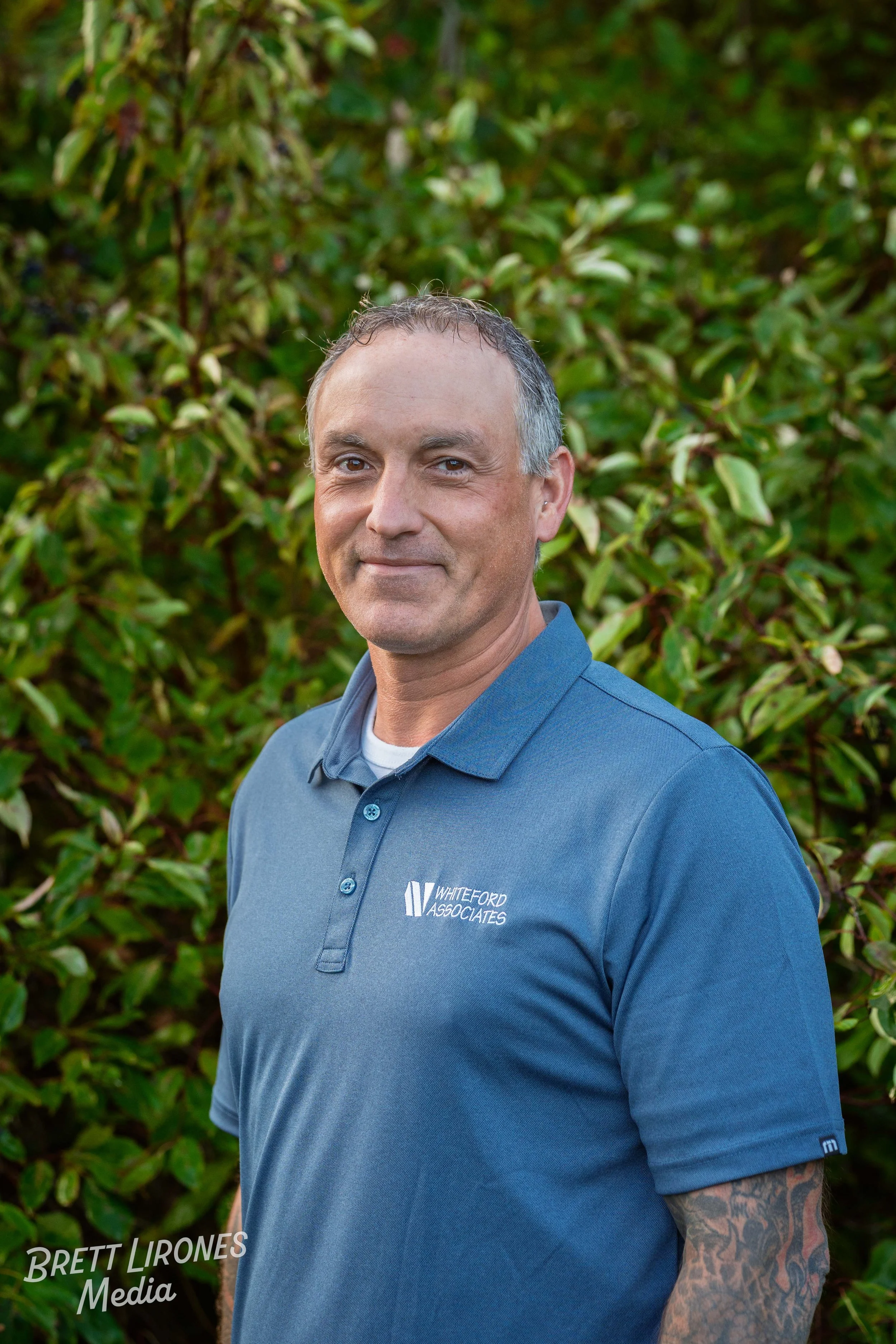 Man with short gray hair wearing a blue polo shirt with 'Whiteford Associates' logo, standing outdoors in front of green shrubbery.
