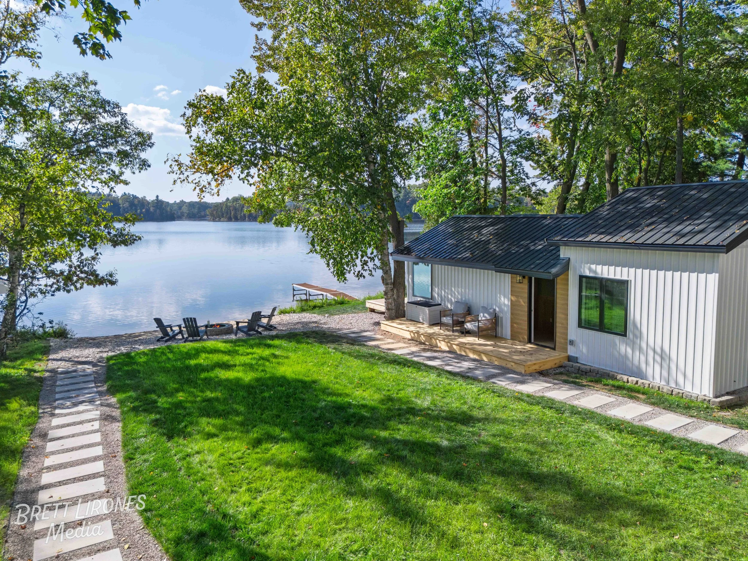 A modern house with white siding and a black metal roof is situated near a lake, surrounded by lush green trees. There is a small deck with outdoor chairs, a gravel pathway, a grassy lawn, and Adirondack chairs around a fire pit by the water.