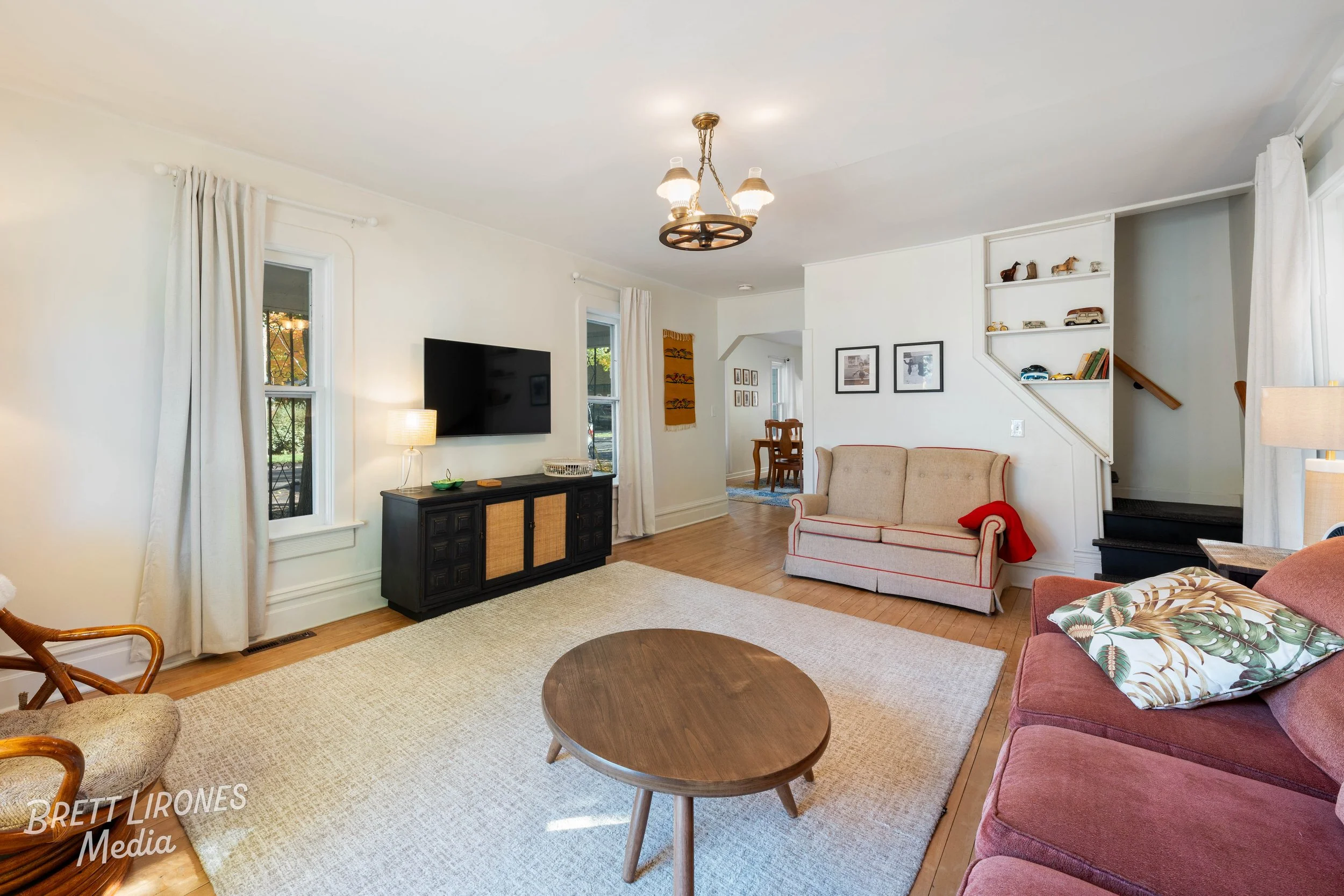 Living room with off-white walls, hardwood floors, a beige rug, a black TV stand, a wall-mounted flat-screen TV, a lamp, a beige sofa with red piping, a pink sofa with decorative pillows, a small round wooden coffee table, a large window with white c