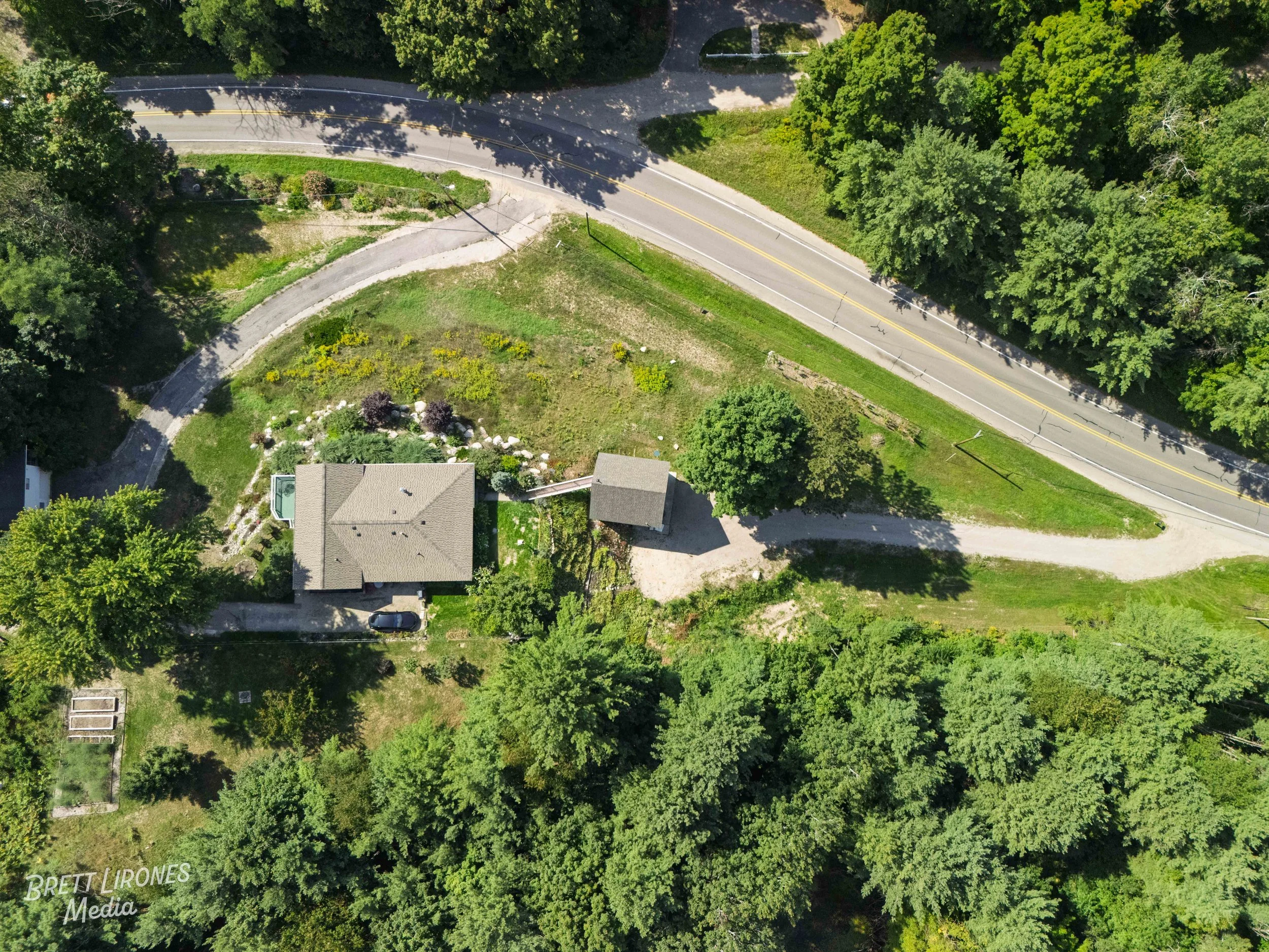 Aerial view of a house surrounded by trees, a curved driveway, and a road with a double yellow line in a green landscape.