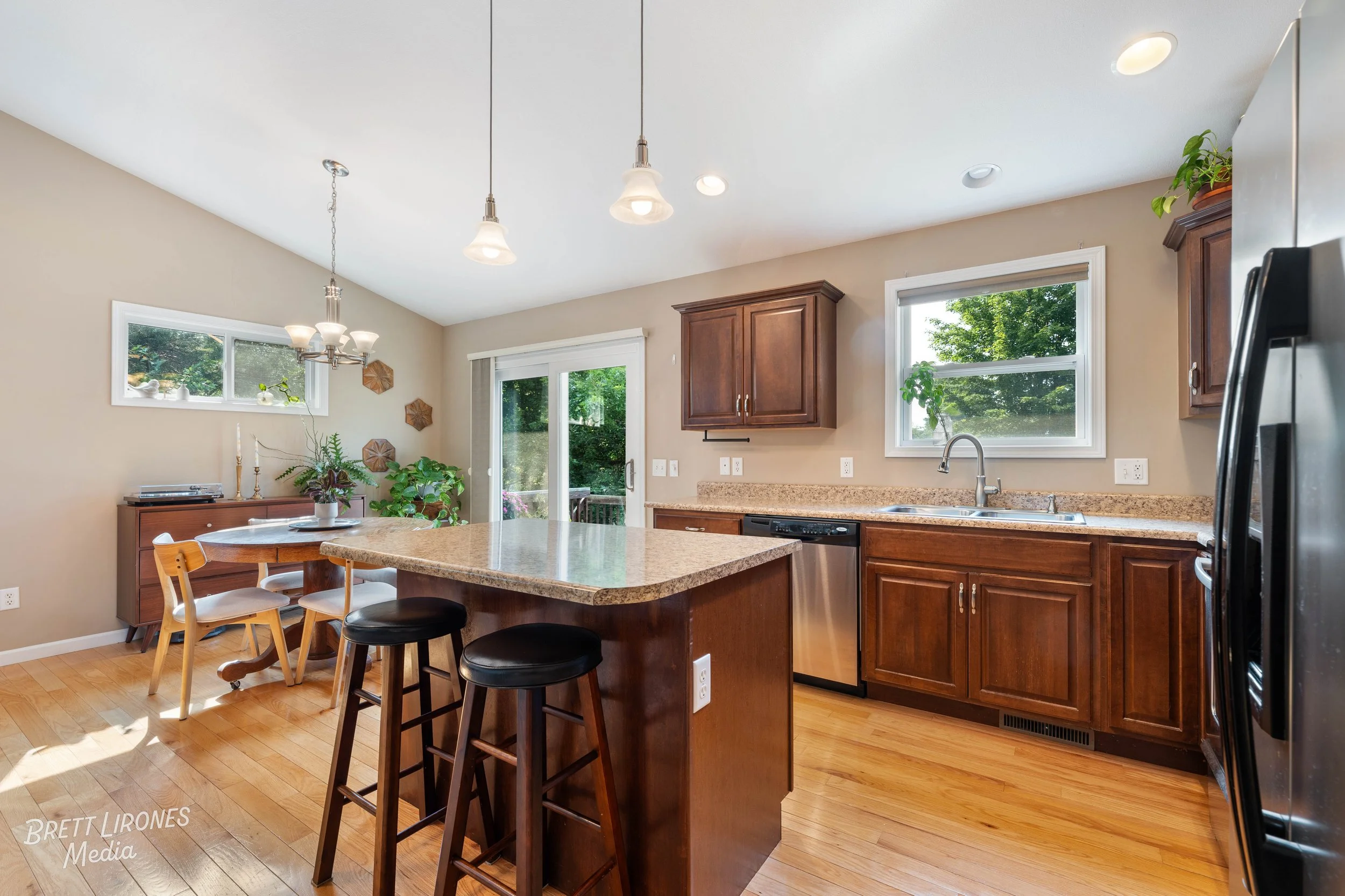 Kitchen with wooden cabinets, beige countertop, stainless steel dishwasher, black refrigerator, double window above sink, dining table with chairs, ceiling lights, hardwood floor, potted plants, glass sliding door leading outside.