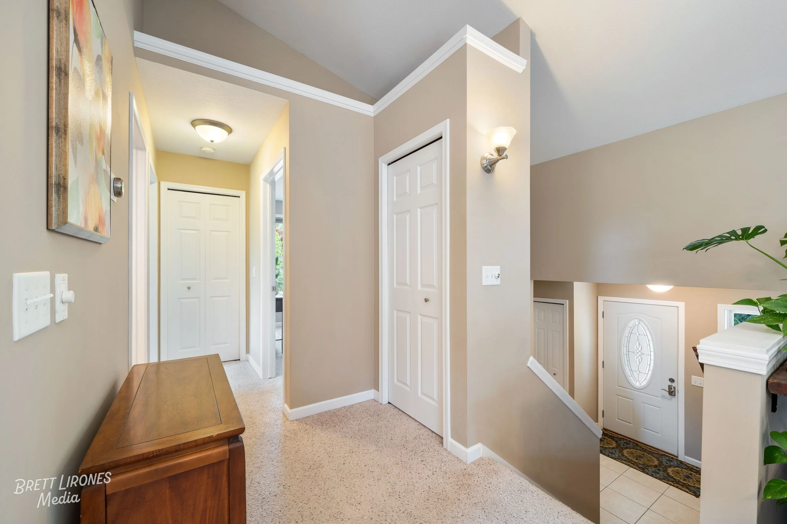 A hallway with beige walls and white trim, displaying a wooden bench, a painting, and multiple closed white doors. There's a staircase leading downstairs with a white front door and decorative window at the bottom.