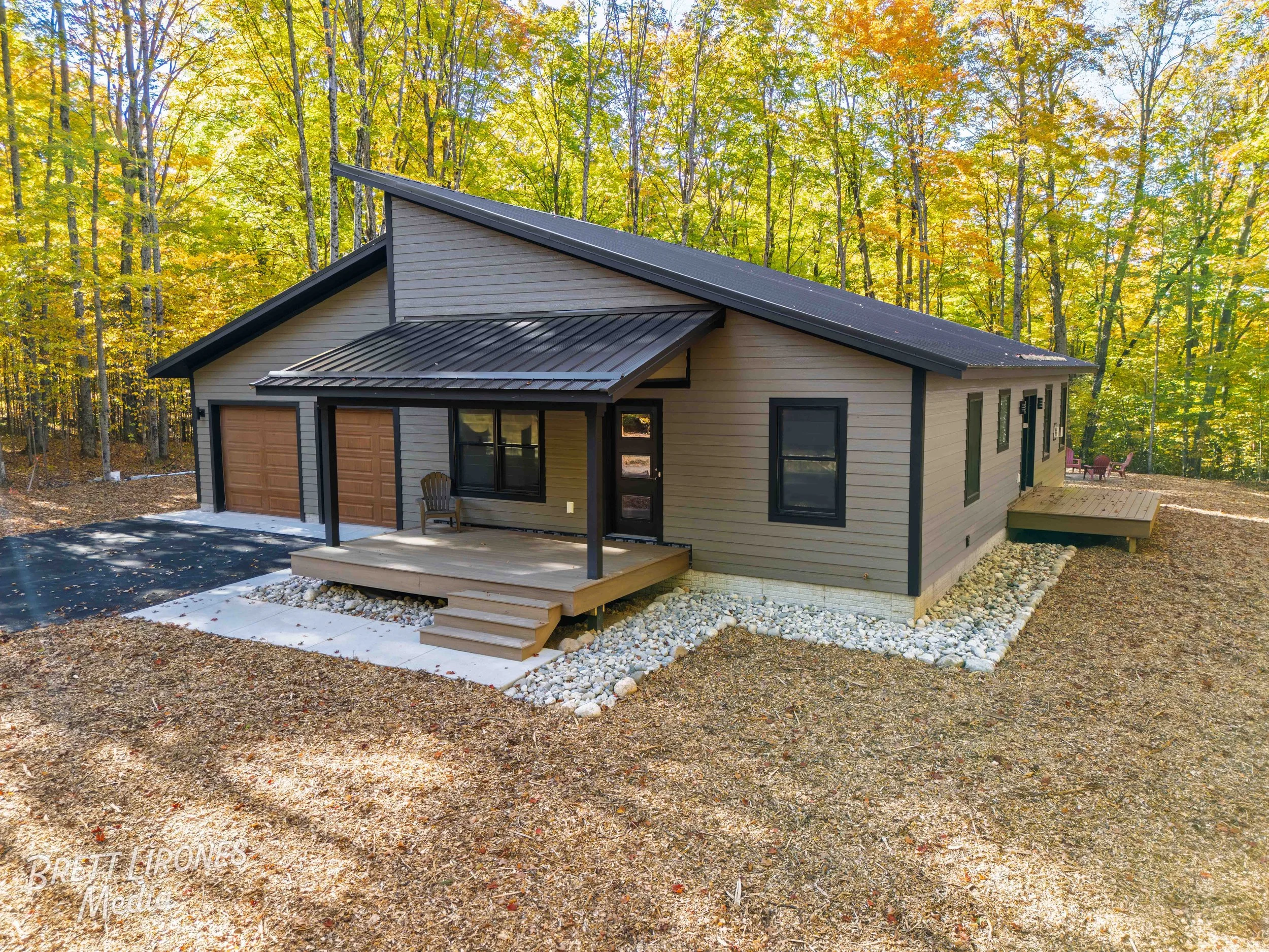 Newly built house in a wooded area with a sloped roof, two garage doors, a front porch with stairs, and outdoor seating, surrounded by trees with autumn foliage.