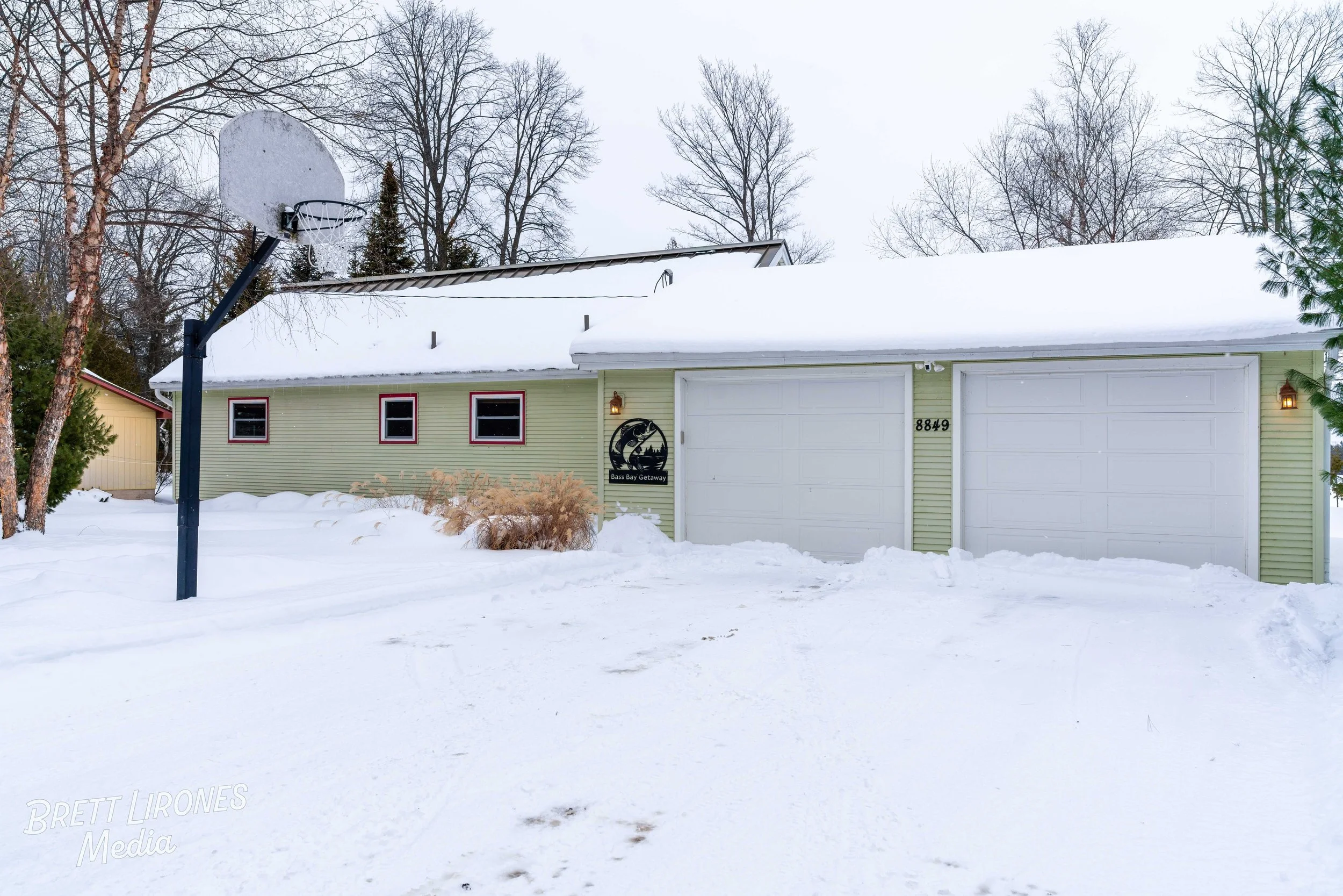 A snow-covered house with a basketball hoop, three red-trimmed windows, and a sign that reads 'Bass Bay Getaway' on the front. The house is pale green with white garage doors and is surrounded by leafless trees in winter.
