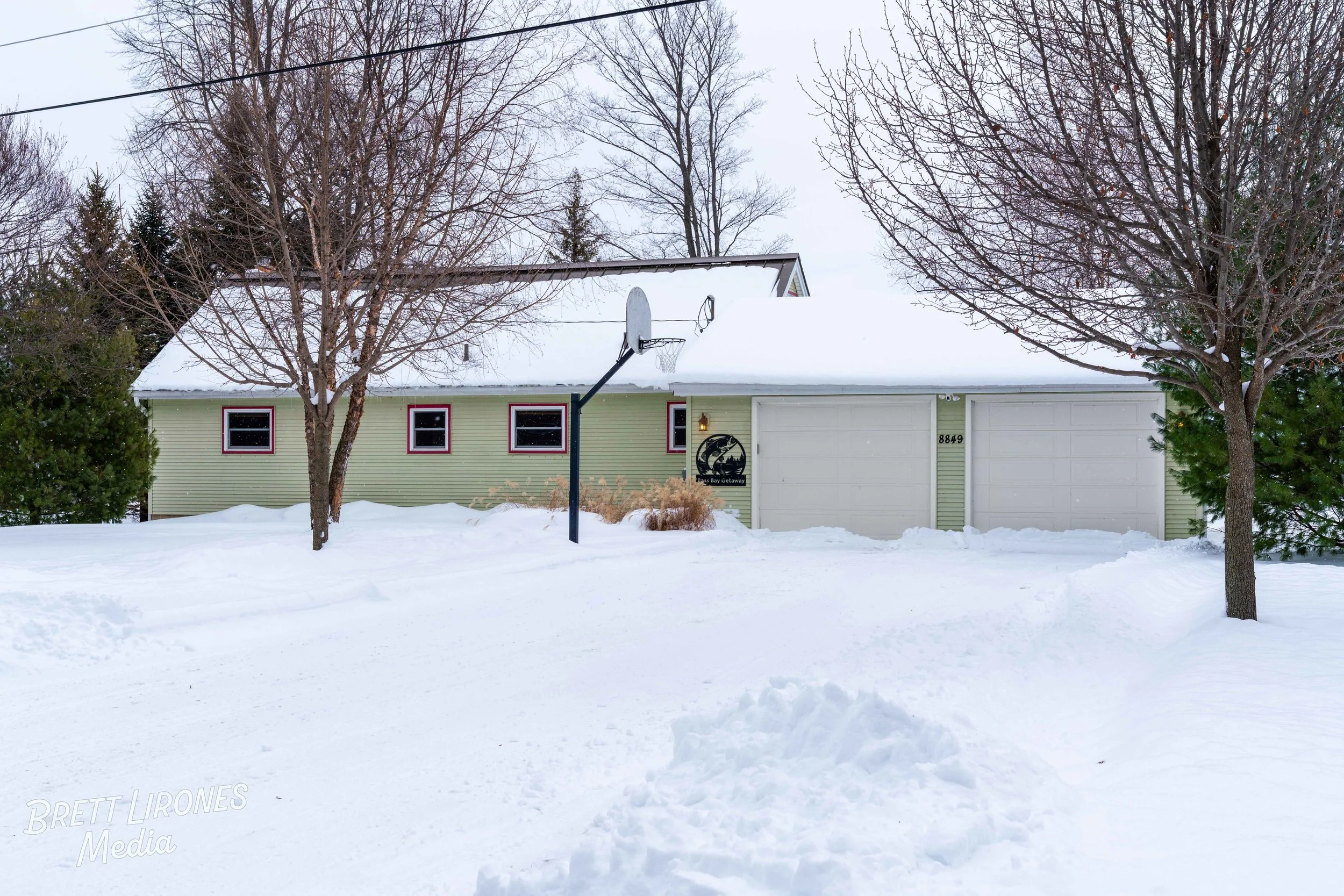 A house with snow-covered yard and driveway, a basketball hoop, leafless trees, and a sign on the house that reads 'Four Bay Gateway.'