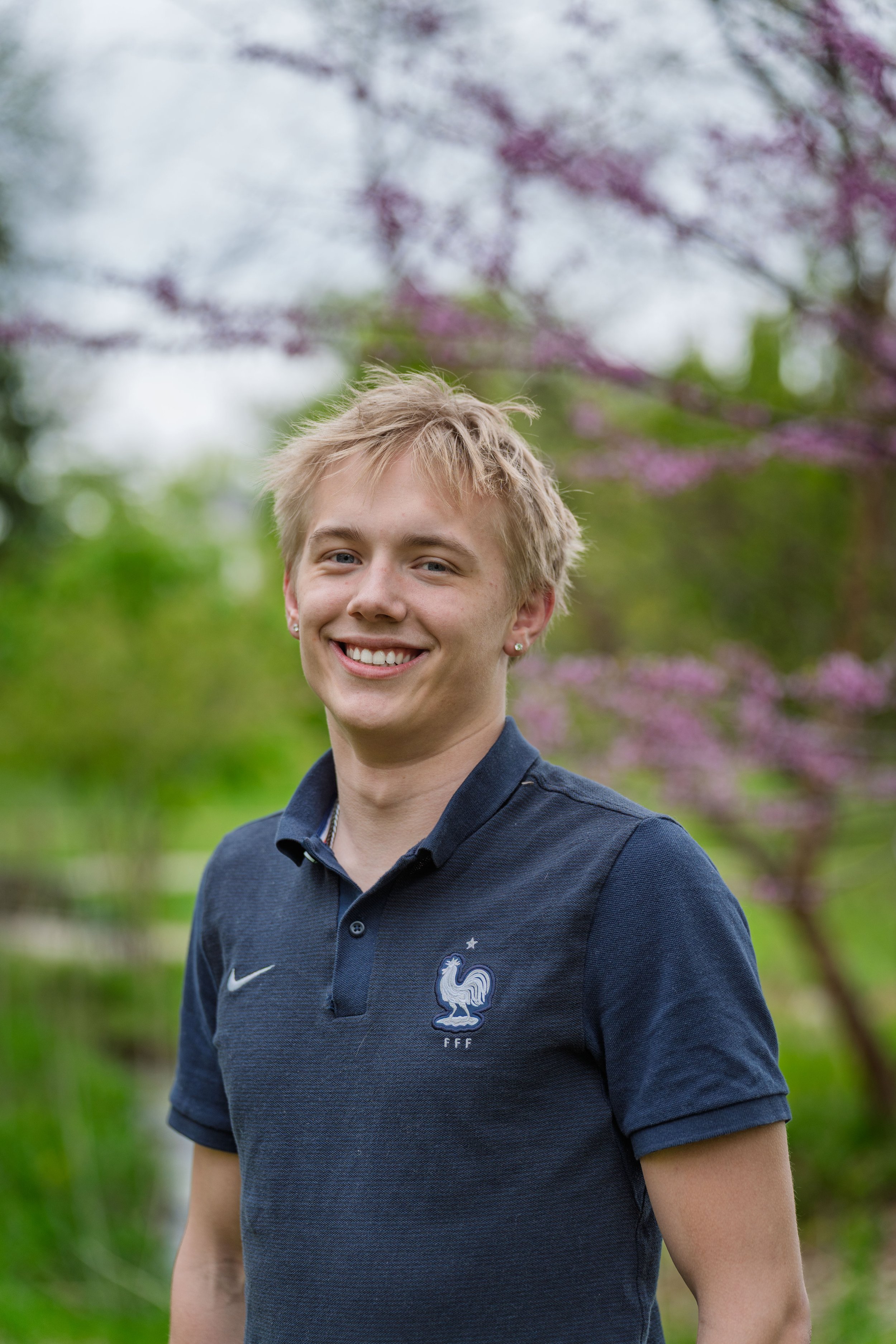 Young man smiling outdoors, wearing a navy blue polo shirt with the French Football Federation logo, in front of blooming trees with pink flowers.