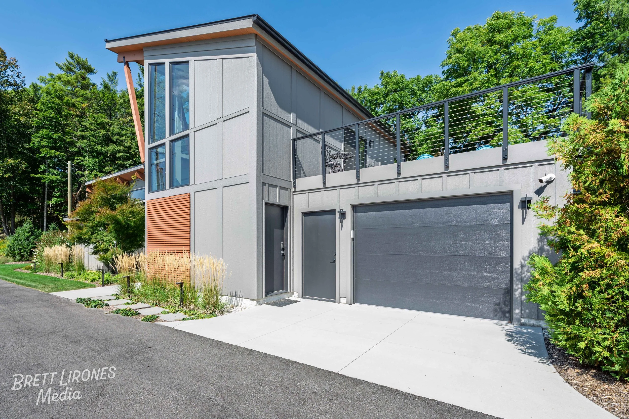 Modern two-story house with large windows, gray exterior, attached garage, and rooftop patio surrounded by greenery under a clear blue sky.