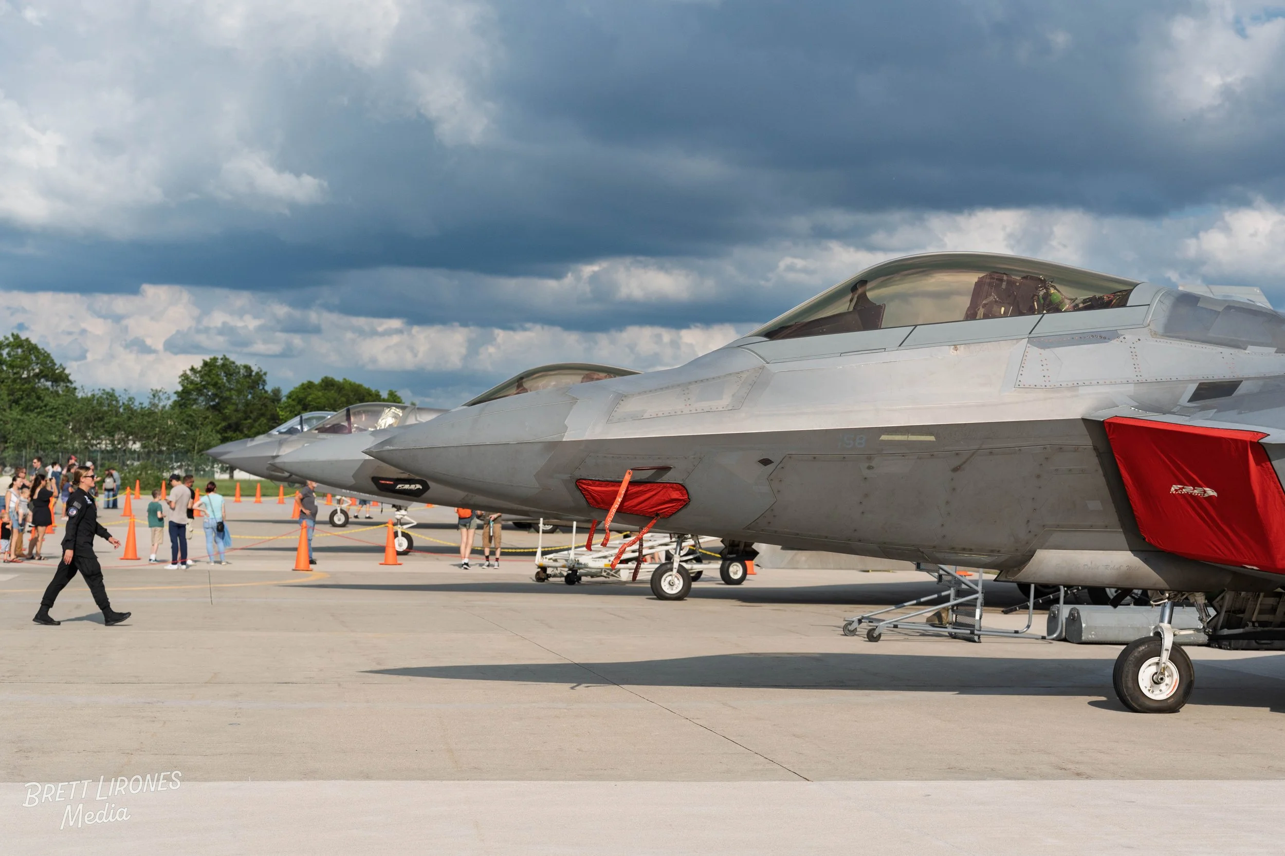 Multiple fighter jets parked on an airfield with people walking around, cones in the background, and a cloudy sky overhead.