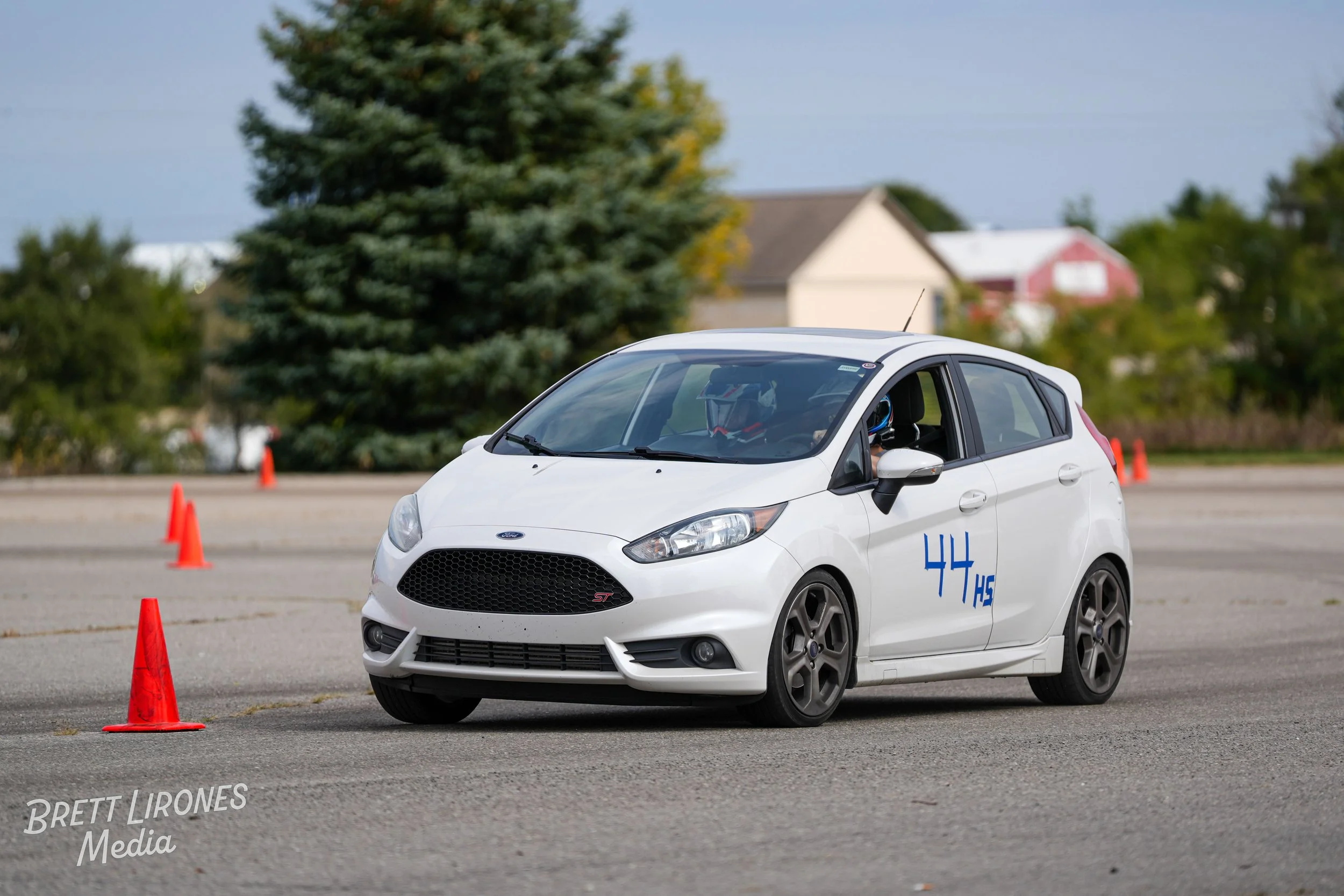 A white Ford Fiesta ST car navigating through an autocross course marked with orange cones, with a driver wearing a helmet visible through the window, on a flat paved surface outdoors with trees and buildings in the background.