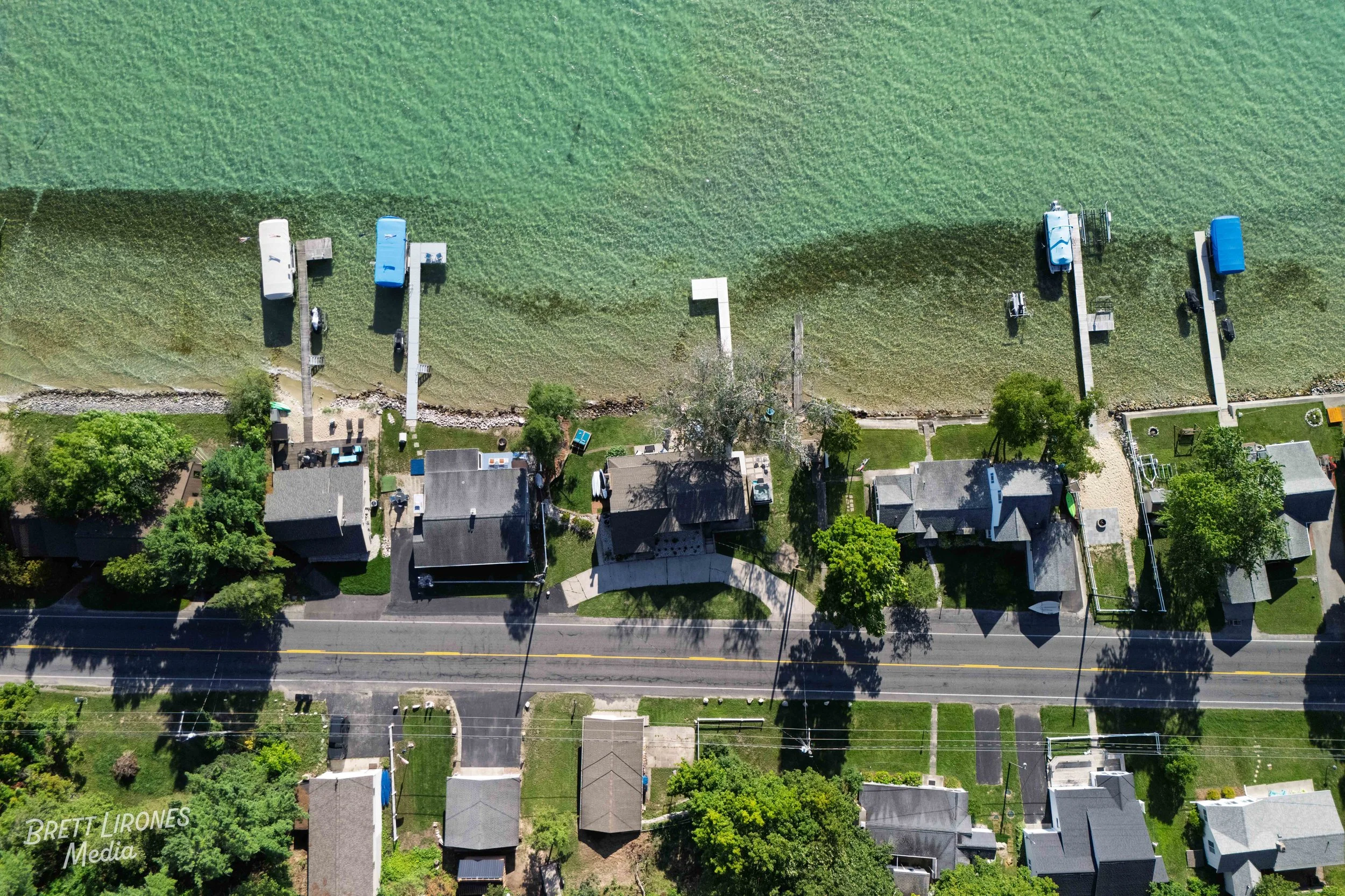 Aerial view of homes along a shoreline with small docks and boats, and a green bay or lake beyond.