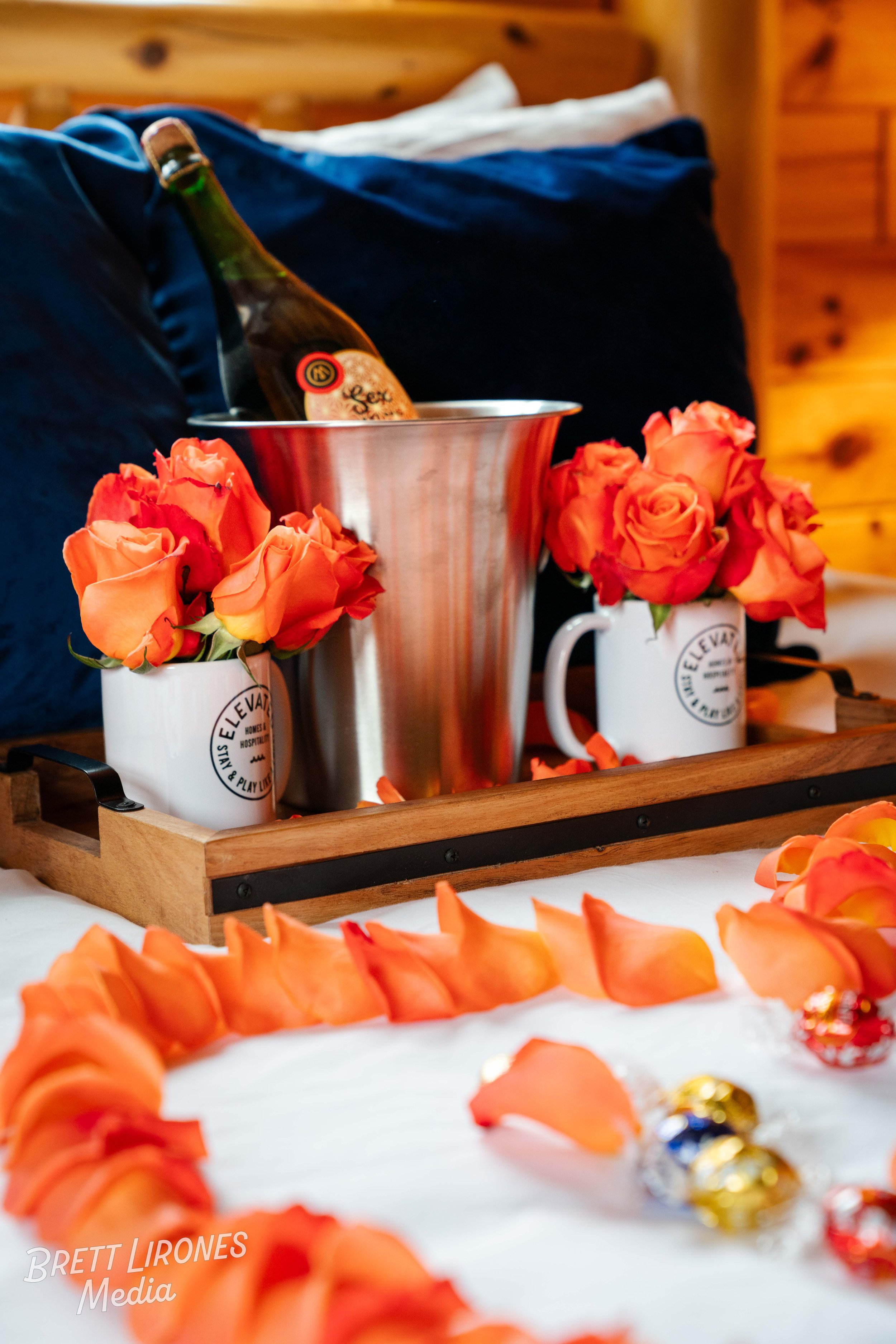 A wooden tray on a bed displays two white mugs with orange roses, a metallic ice bucket with a bottle, and a decorative arrangement of orange rose petals on the bedspread.