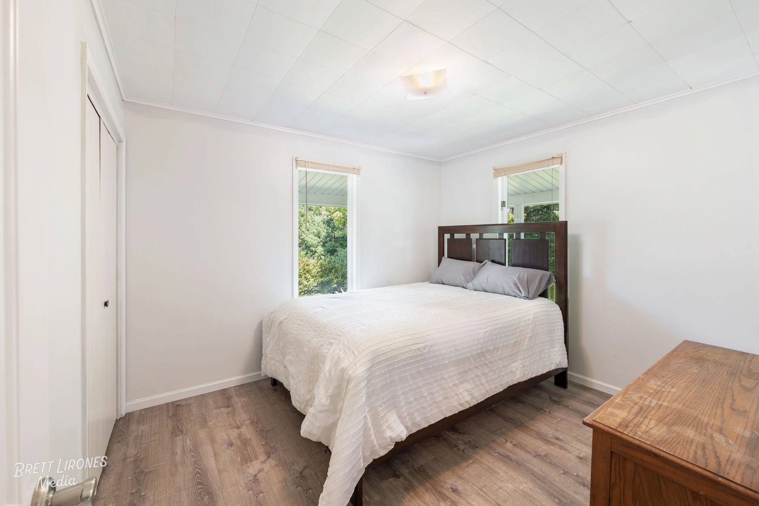 A bedroom with a bed, two windows with blinds, a white bedspread, gray pillows, a wooden dresser, and hardwood floors.