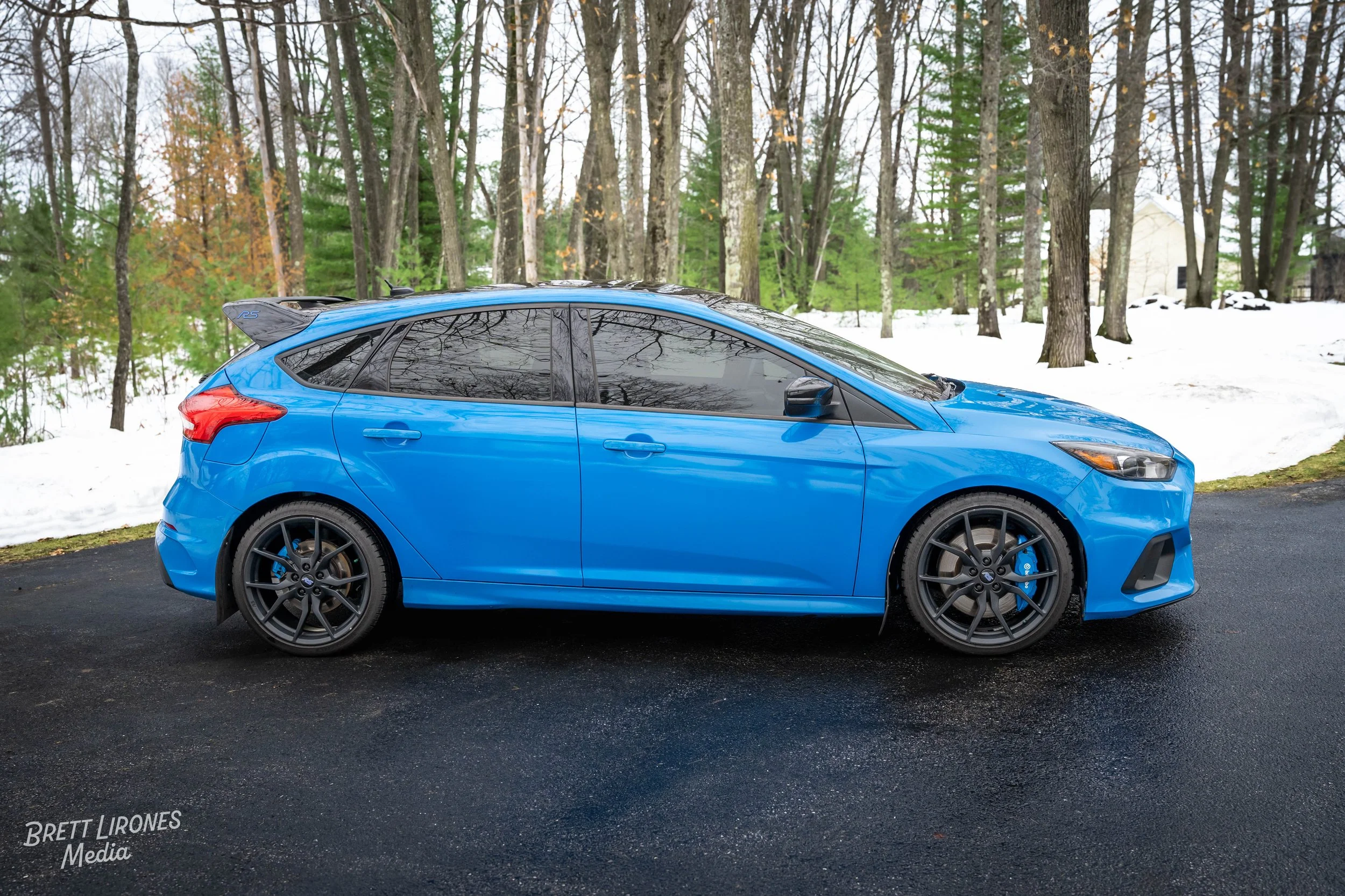 Blue hatchback car parked on a dark asphalt driveway with snow and trees in the background.