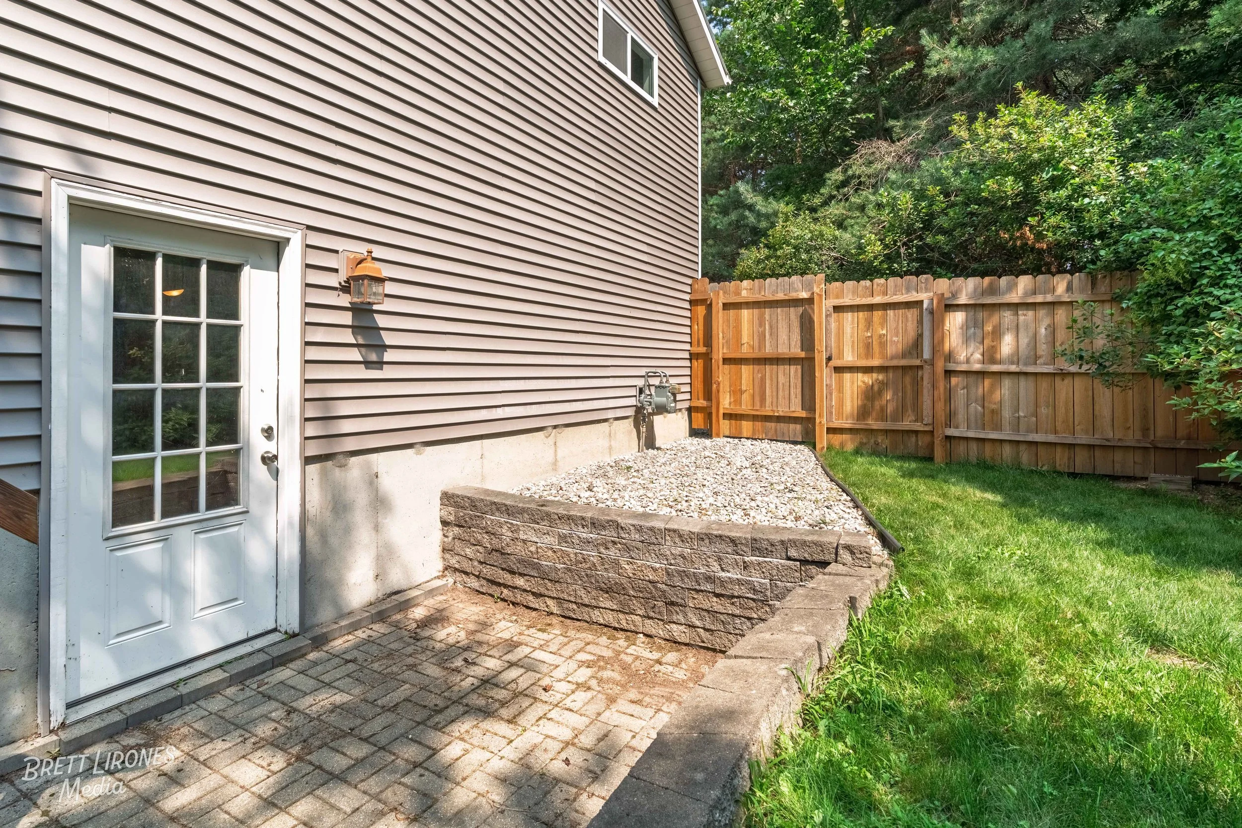 Backyard corner with a gray house wall, white door with glass panes, a wall-mounted outdoor light, a gravel bed bordered by a brick retaining wall, green grass, and a wooden privacy fence with trees behind.
