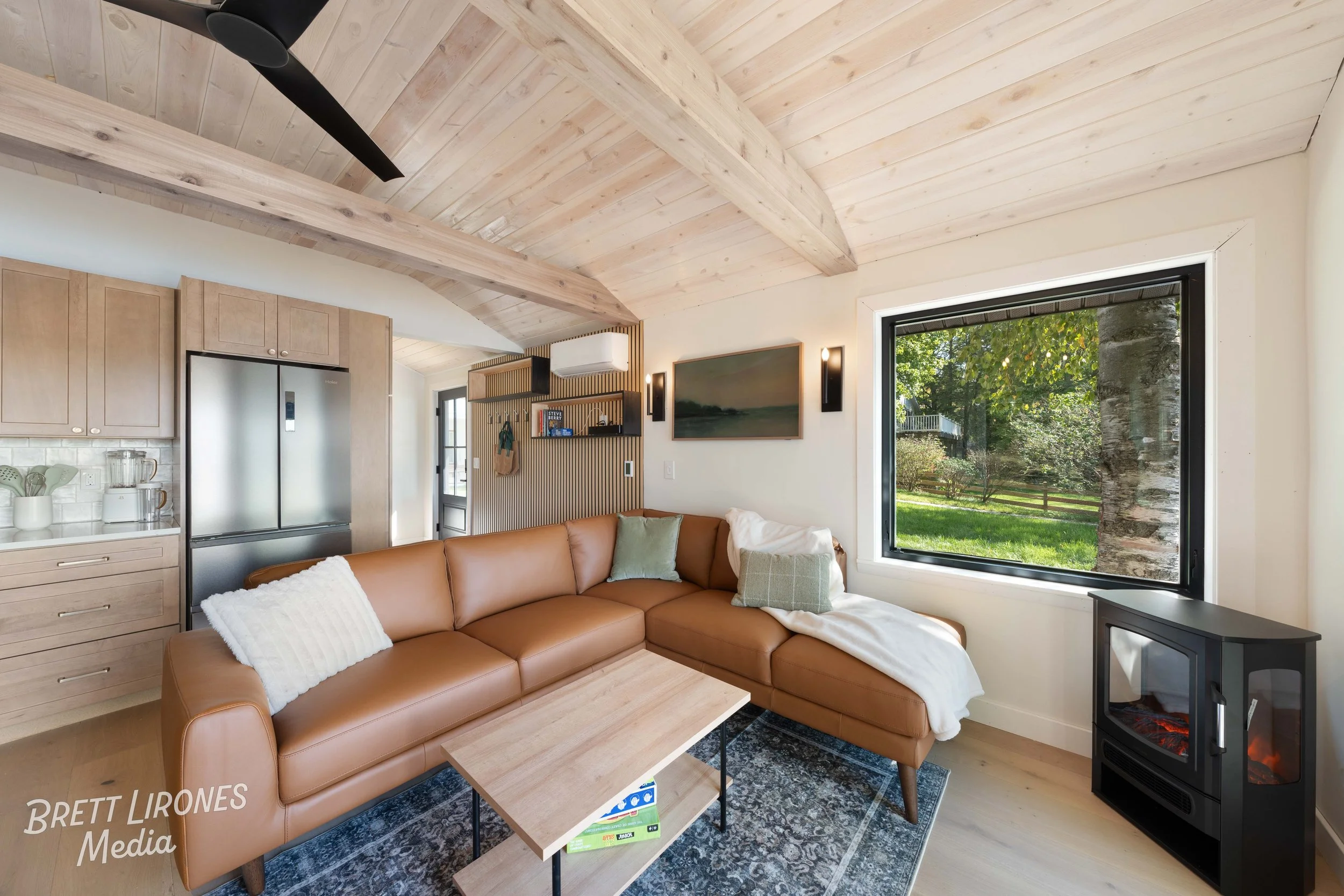 Living room with a brown leather sectional sofa, a small wooden coffee table, a window showing a green outdoor space, and a black electric fireplace.