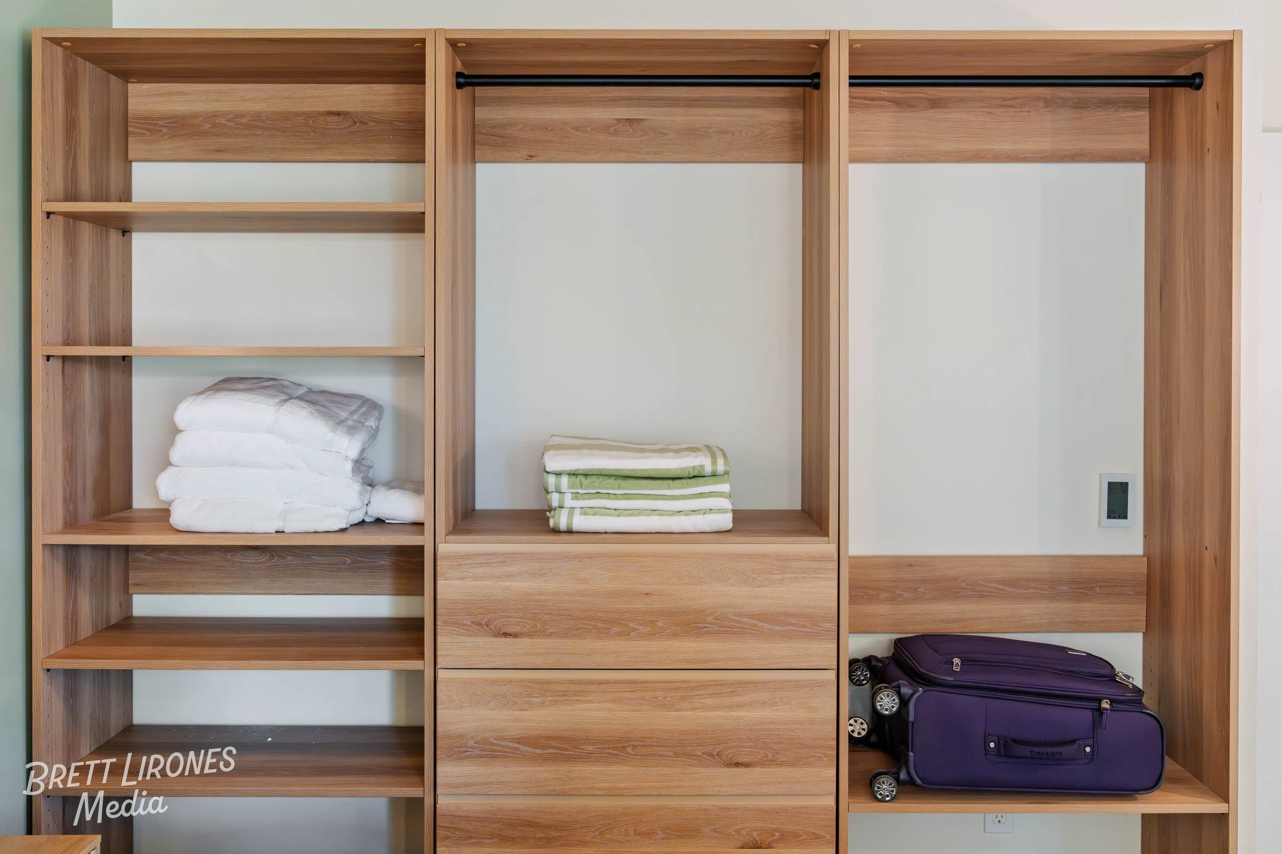 Open wooden closet with shelves, folded towels, a suitcase, and a wall-mounted thermostat