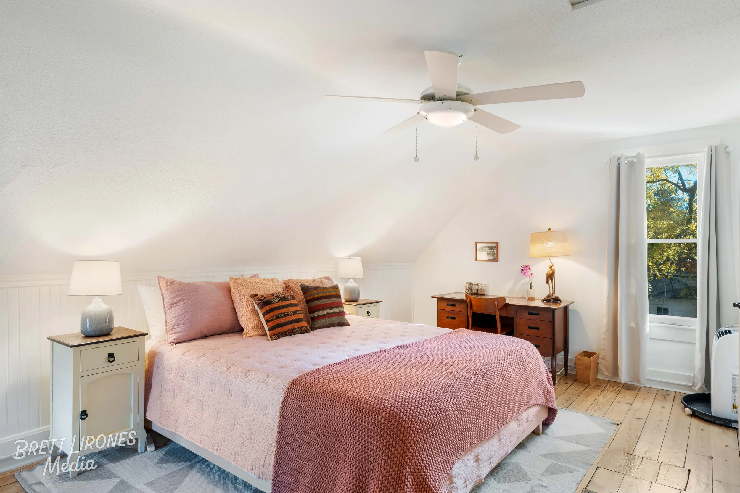 A cozy bedroom with a large bed covered in pink bedding and multiple pillows, two nightstands with lamps, a wooden desk with a lamp and flower vase, a window with white curtains showing greenery outside, and a ceiling fan.
