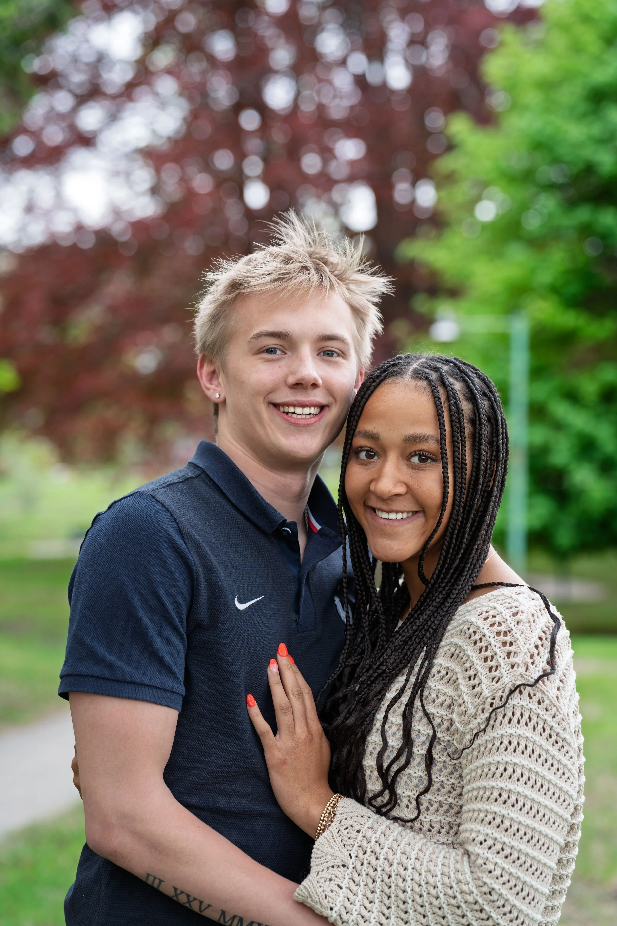 Smiling young couple embracing outdoors in a park with green trees and red foliage in the background.