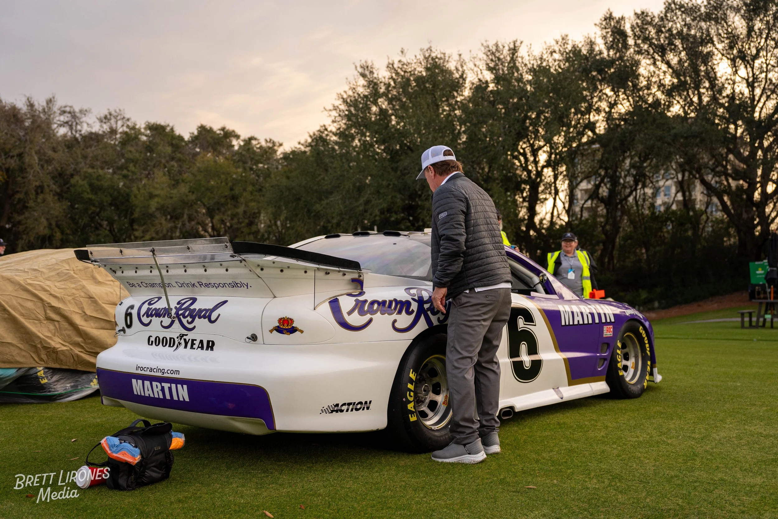 A person in a gray jacket and gray pants standing next to a white and purple race car on a grassy field, with two people in yellow vests in the background and trees behind them at sunset.