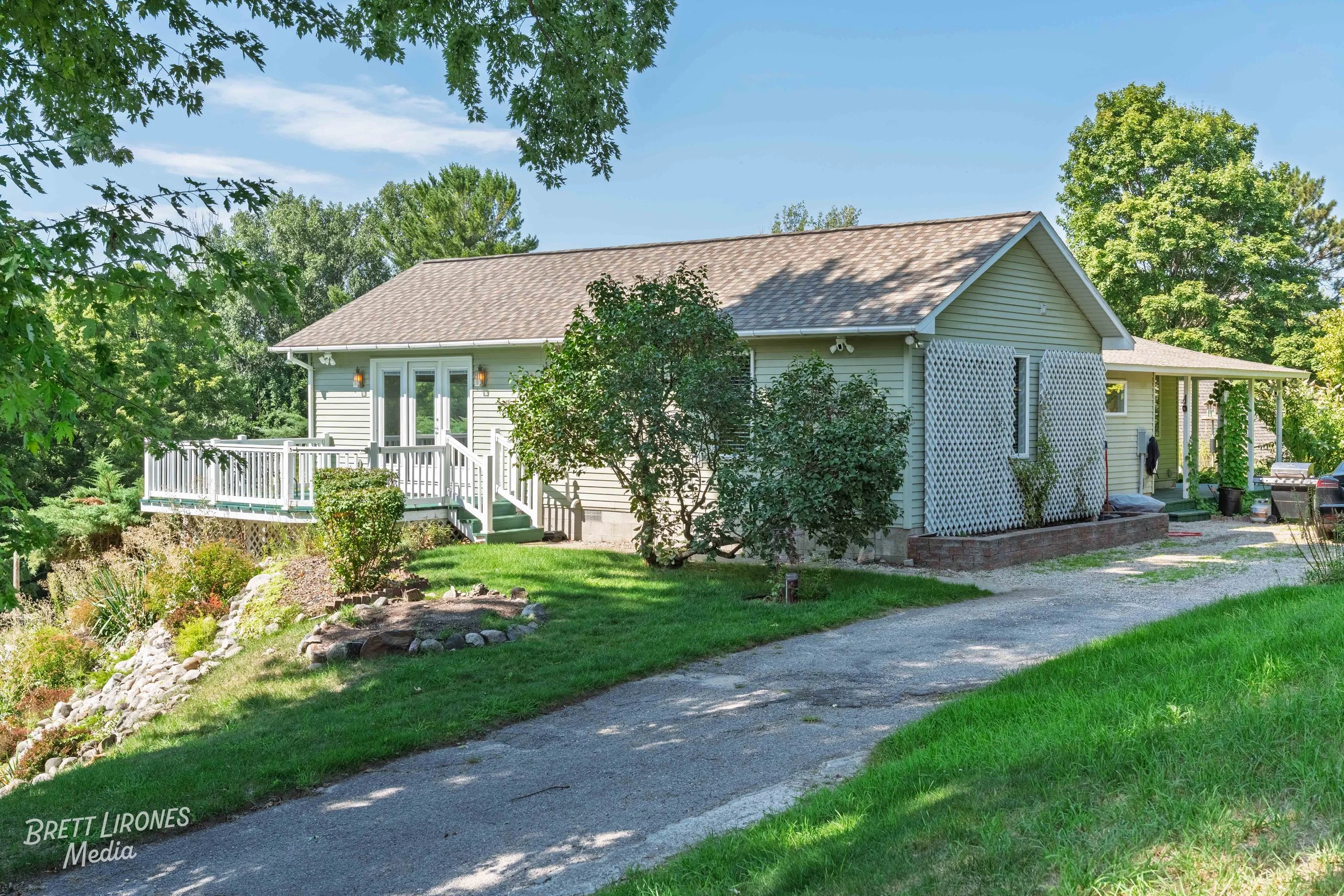 A cozy single-story house with light green siding, a front porch with white railing, surrounded by greenery and trees on a bright, sunny day.