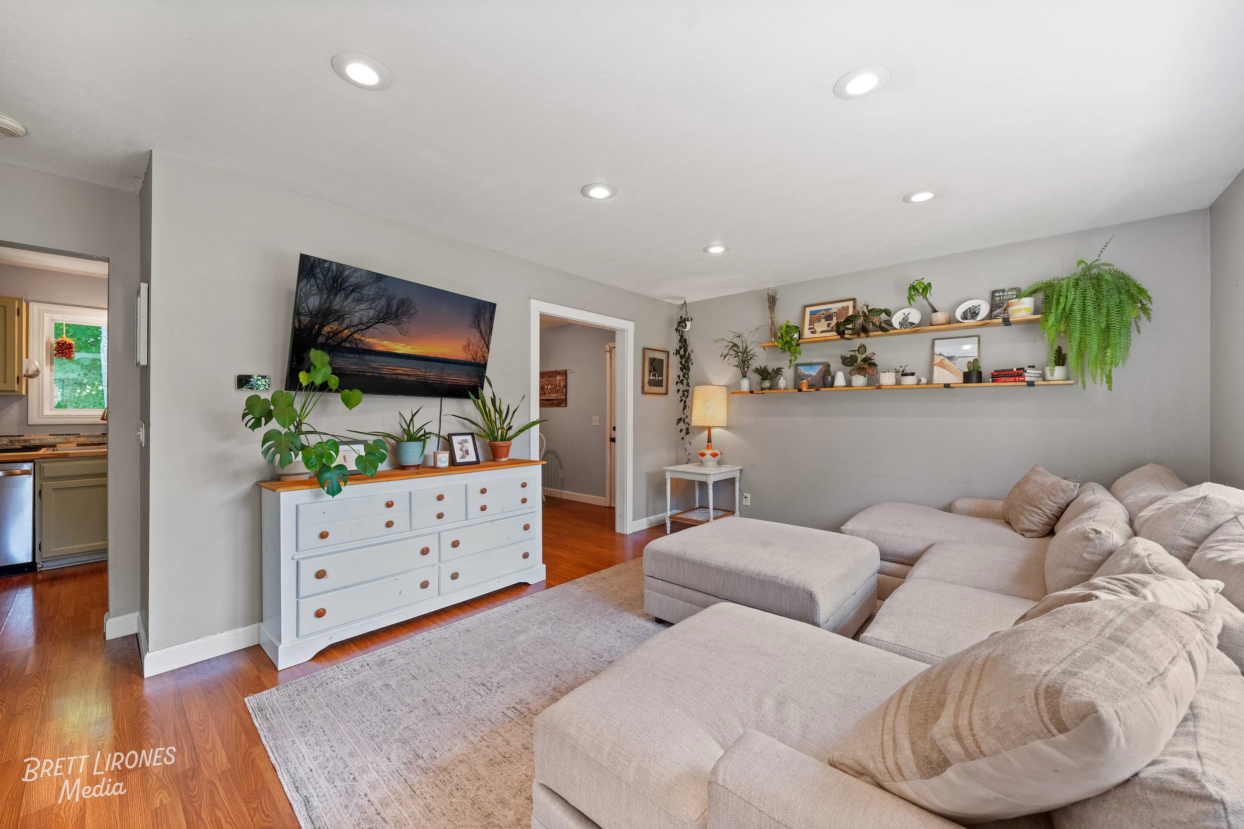 Living room with a beige sectional sofa, potted plants on a white dresser beneath a mounted flat-screen TV, wooden floating shelves with decorative items and plants, a side table with a lamp, and a doorway leading to another room.