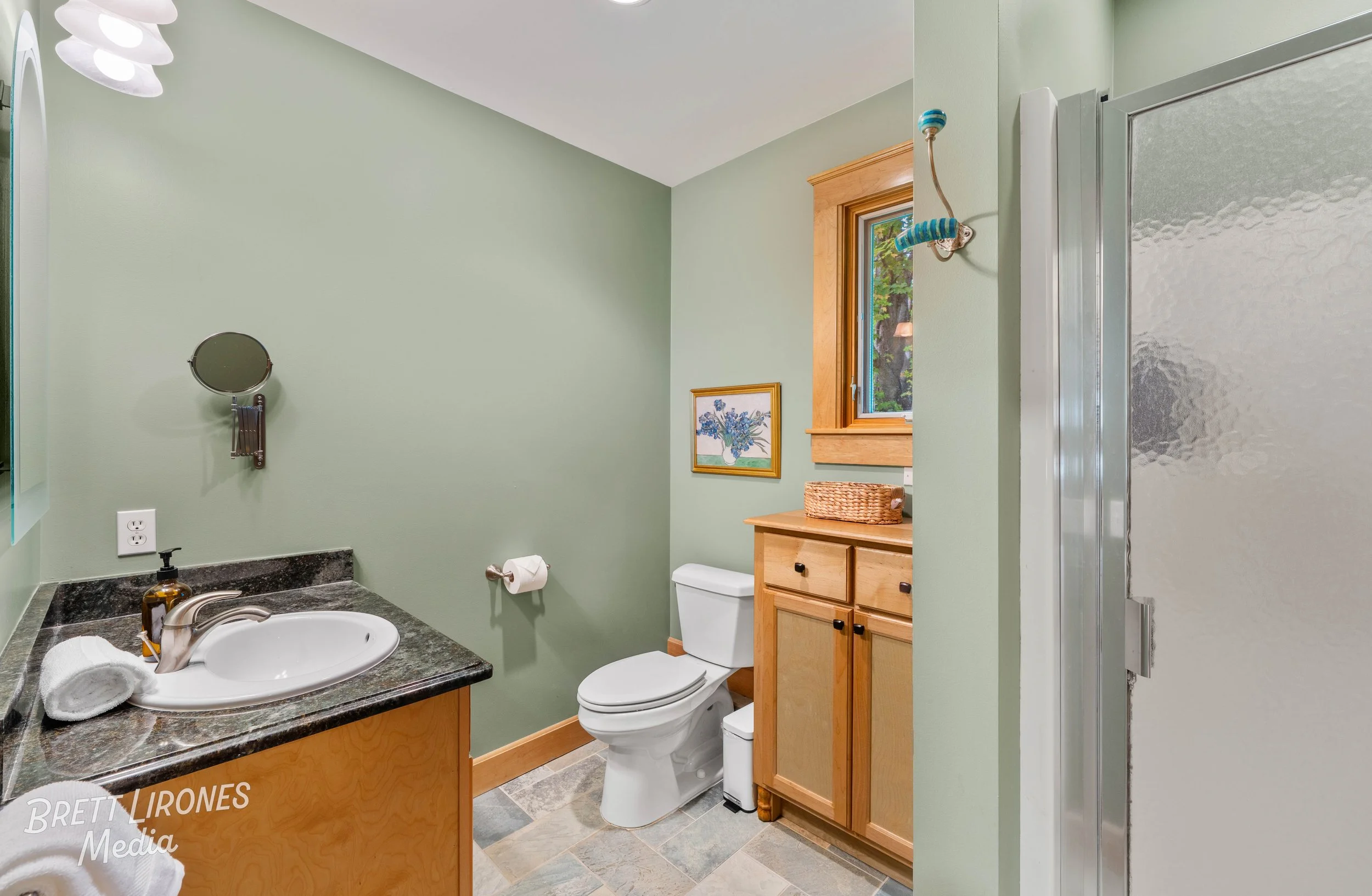 A bathroom with green walls, wooden accents, a sink with a soap dispenser and a rolled towel, a toilet, a wooden cabinet with a basket on top, a window with a view of greenery, a framed artwork, and a textured shower door.