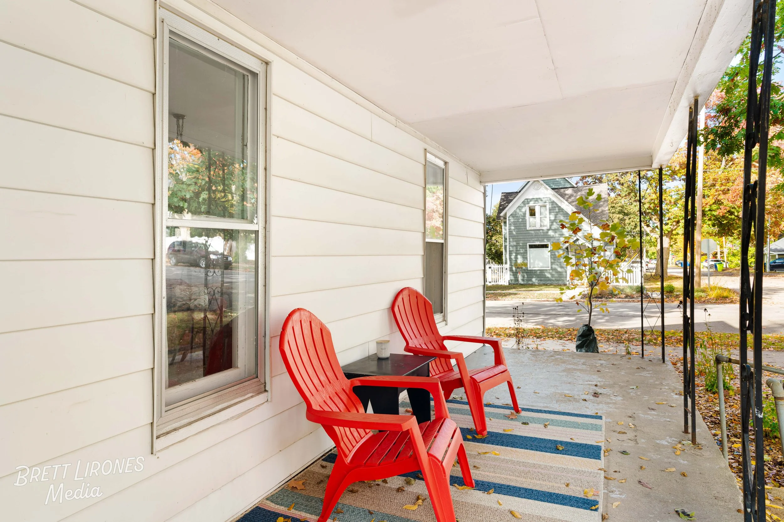 A porch with two red plastic chairs, a small black table, a striped rug, white siding, and a view of a street with trees and houses in the background.