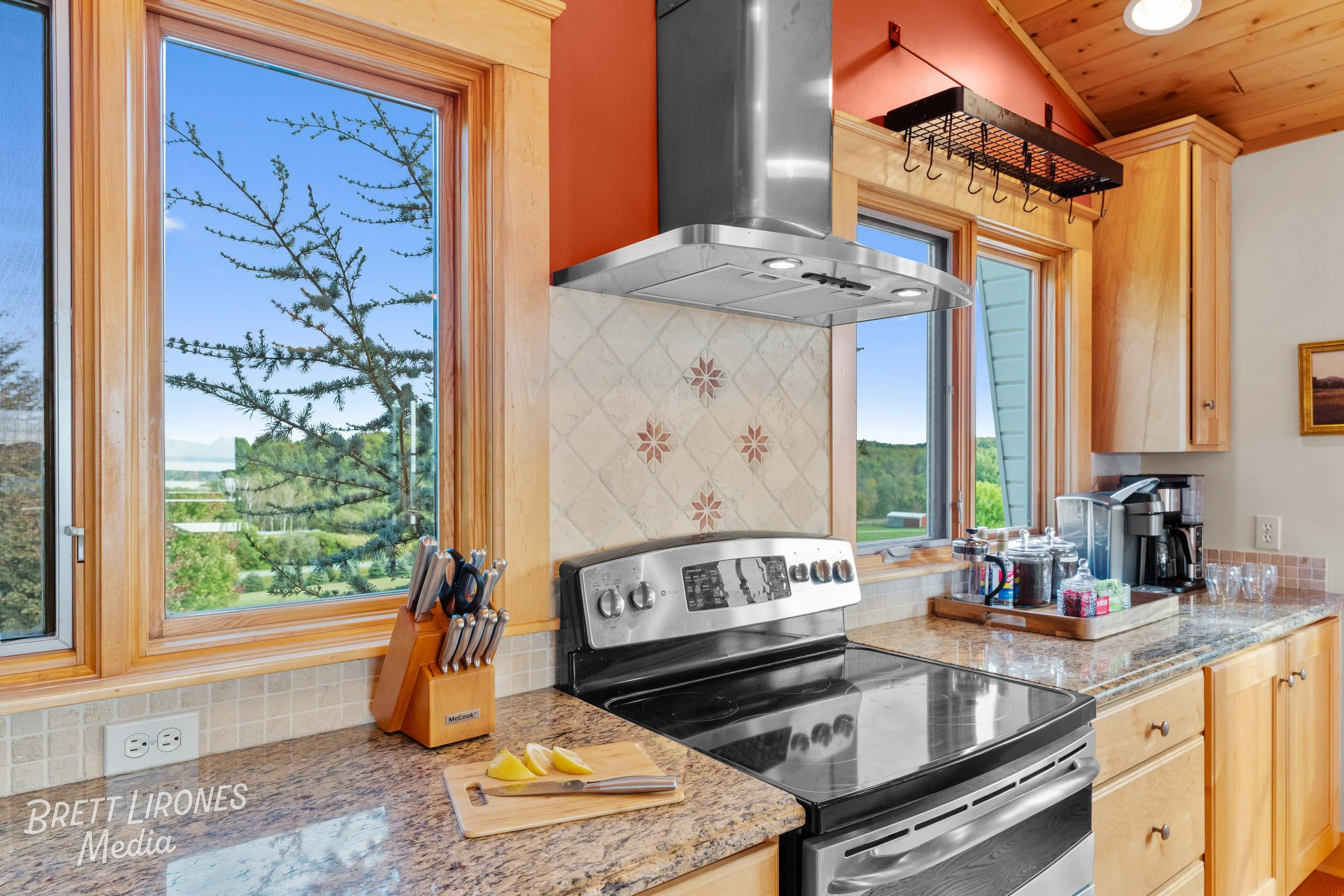 Kitchen with wooden cabinetry, granite countertops, stainless steel stove with vent hood, windows showing outdoor scenery, knife set, lemons, coffee maker, and various kitchen items.