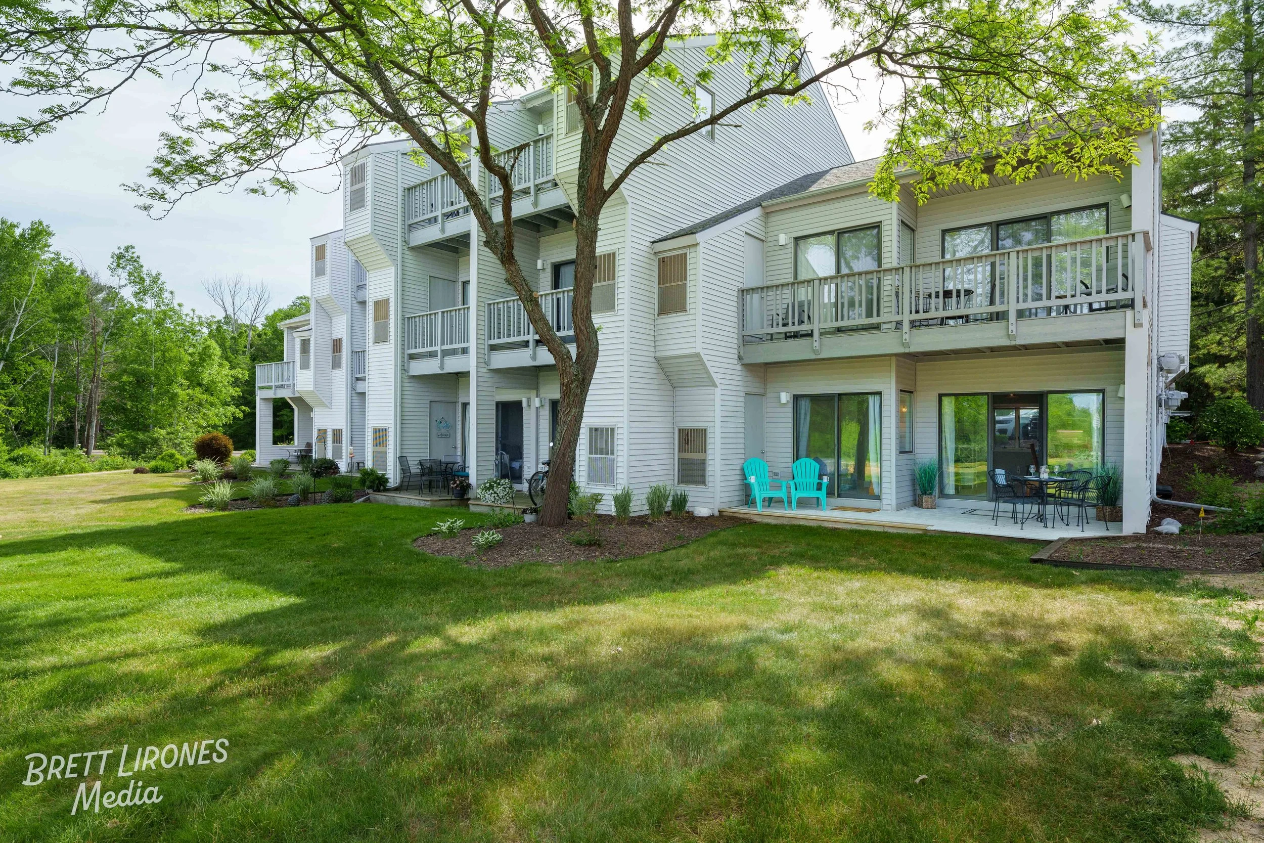 Rear view of a multi-story white apartment building with balconies, large windows, and a well-kept lawn with outdoor furniture, trees, and greenery surrounding it.