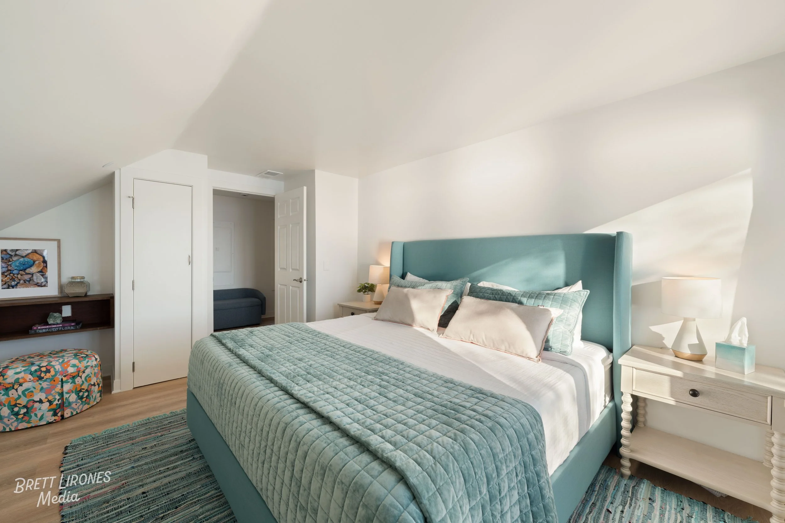 A neatly made bed with teal headboard, several pillows, and a quilted teal bedspread in a bedroom with white walls, two bedside tables with lamps, and a small colorful bench at the foot of the bed.