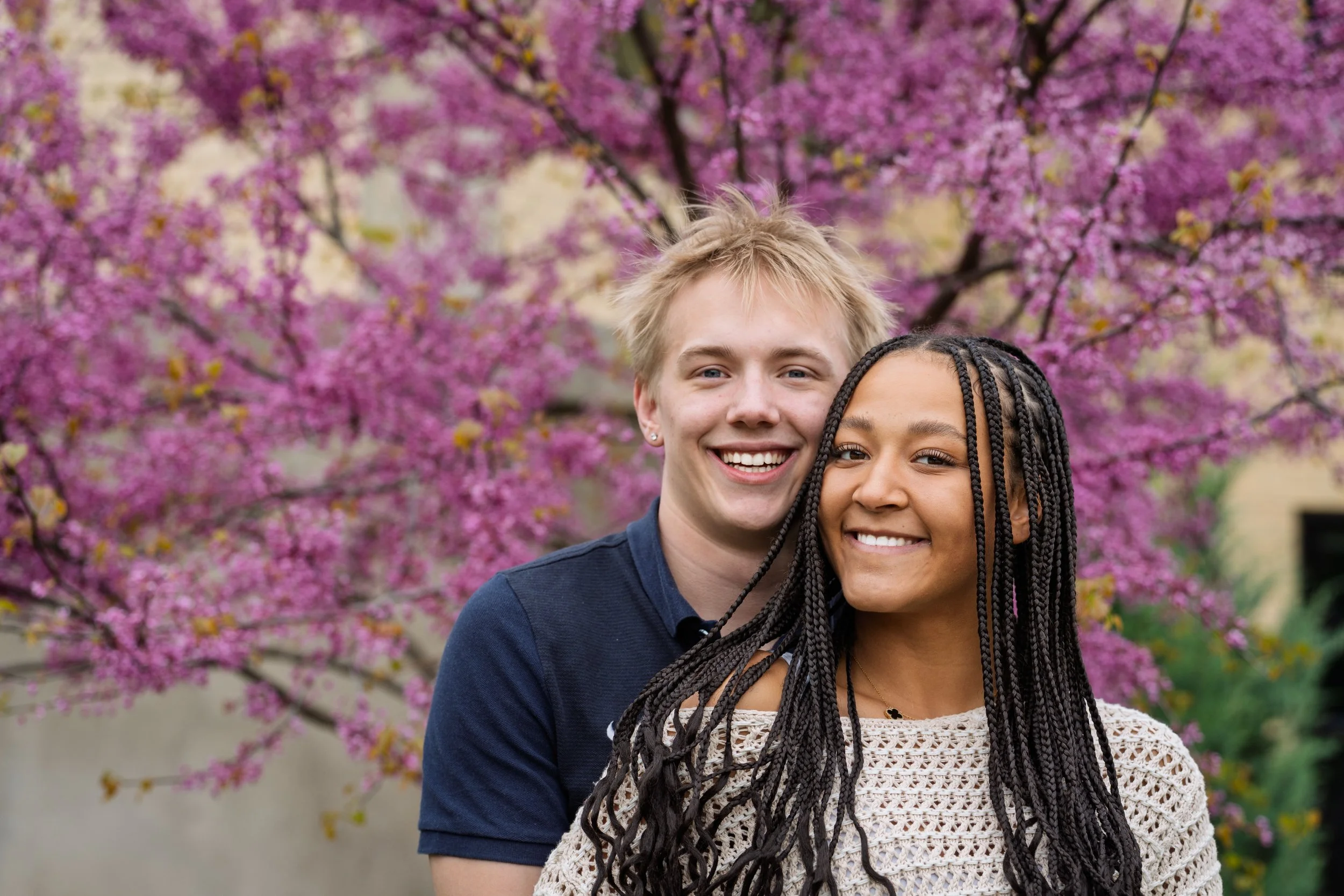 A smiling young man with blonde hair and earrings standing behind a smiling young woman with braided hair, both outdoors in front of a blooming pink tree.