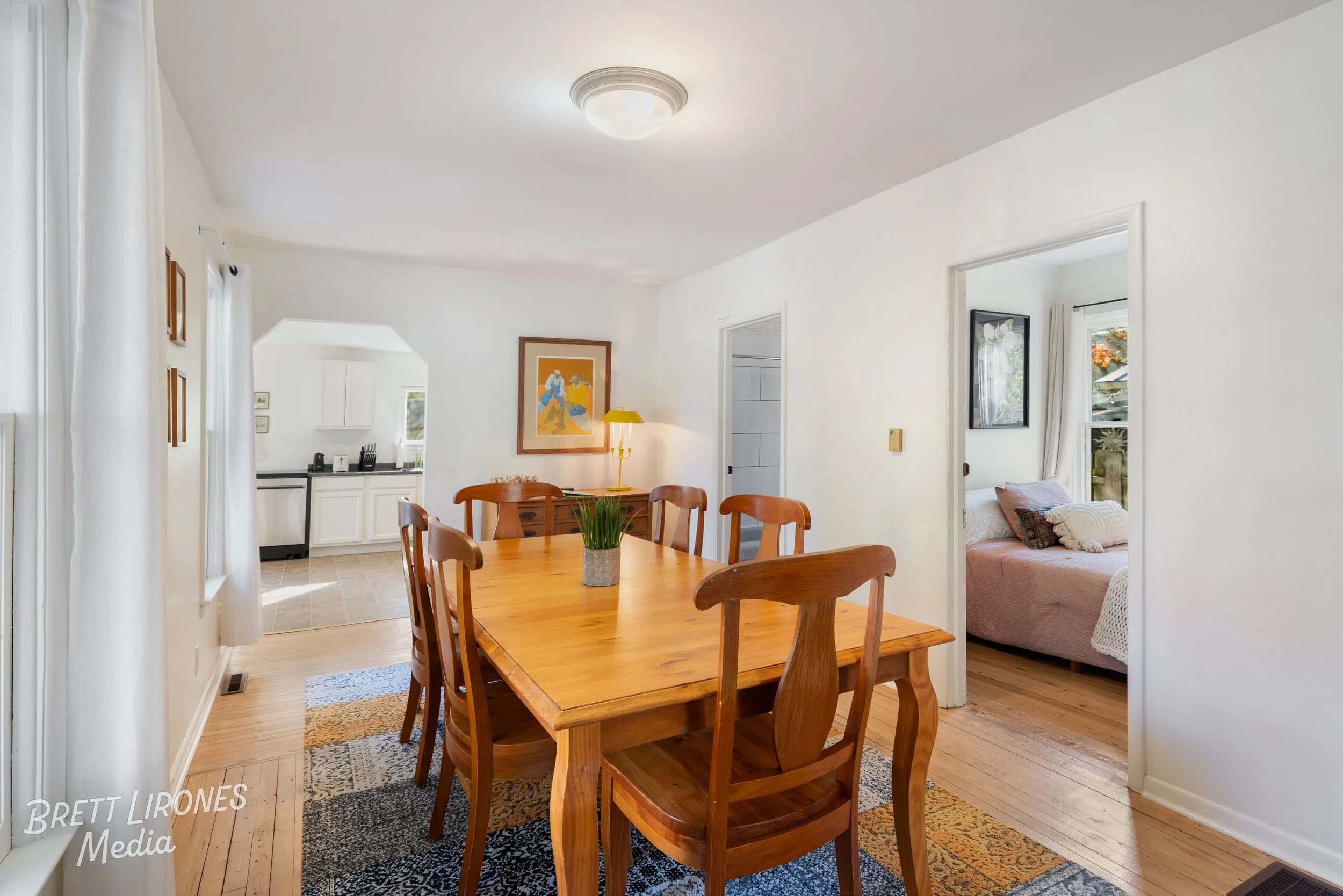 Dining room with wooden table and chairs, a potted plant centerpiece, and abstract artwork on the wall. Visible kitchen through an open doorway, and a bedroom with a bed and window in the background.