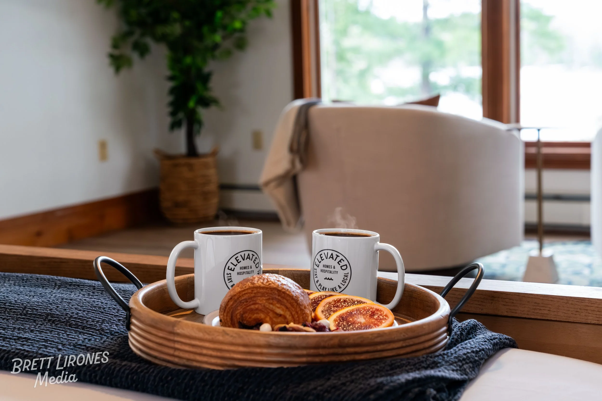 Two white coffee mugs with black text on a wooden tray, accompanied by croissants and dried orange slices on a dark cloth, with a blurred cozy living room in the background featuring a large window, a beige armchair, and a leafy potted plant.