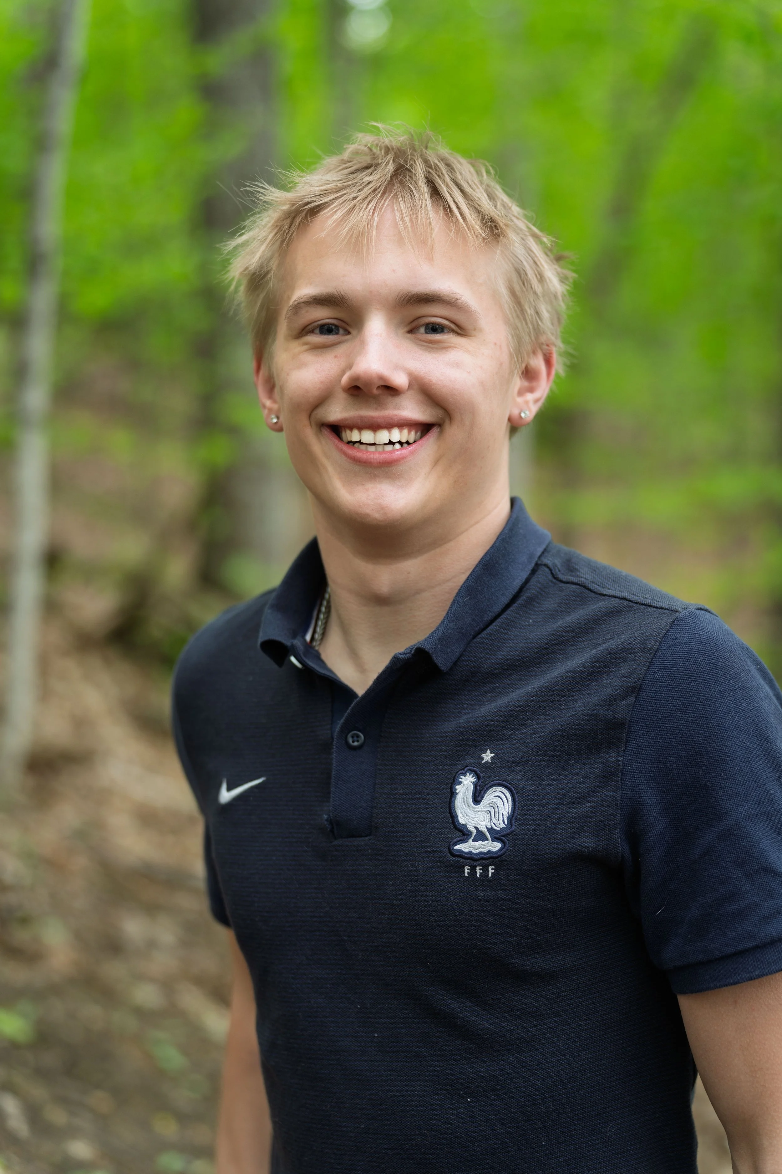 A young man with blonde hair smiling outdoors, wearing a navy blue polo shirt with the France national football team logo.