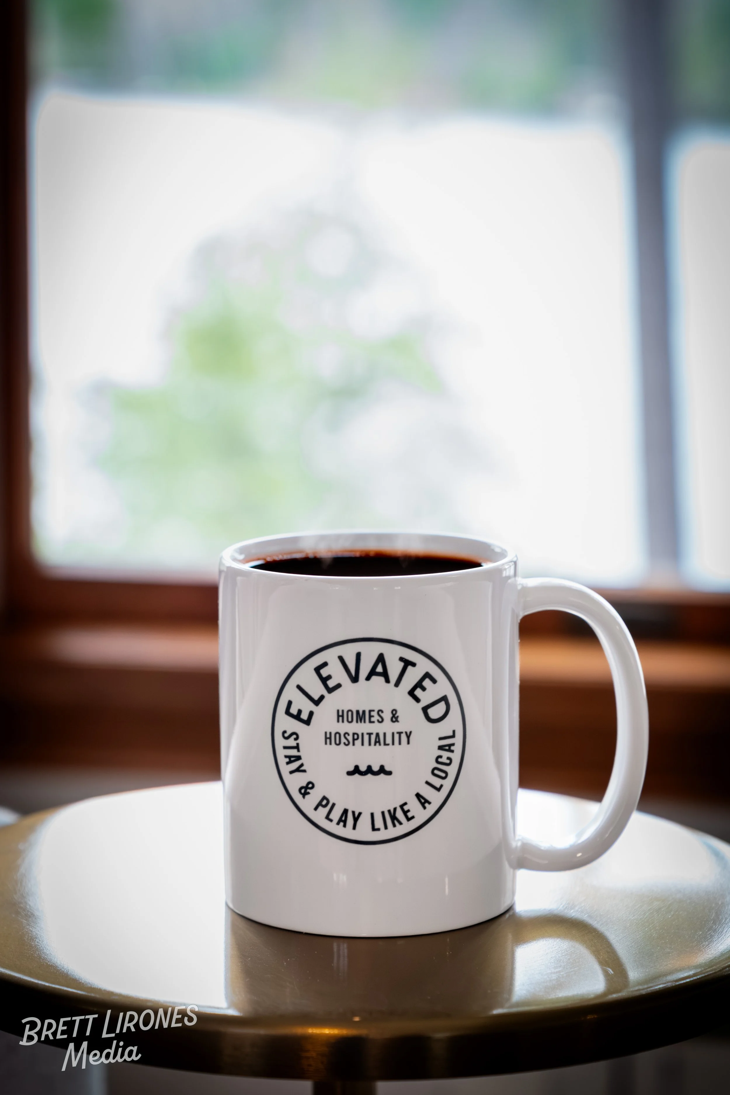 White coffee mug on a tray with a logo that reads "Elevated" and "Homes & Hospitality." The background shows a blurred window with greenery outside.