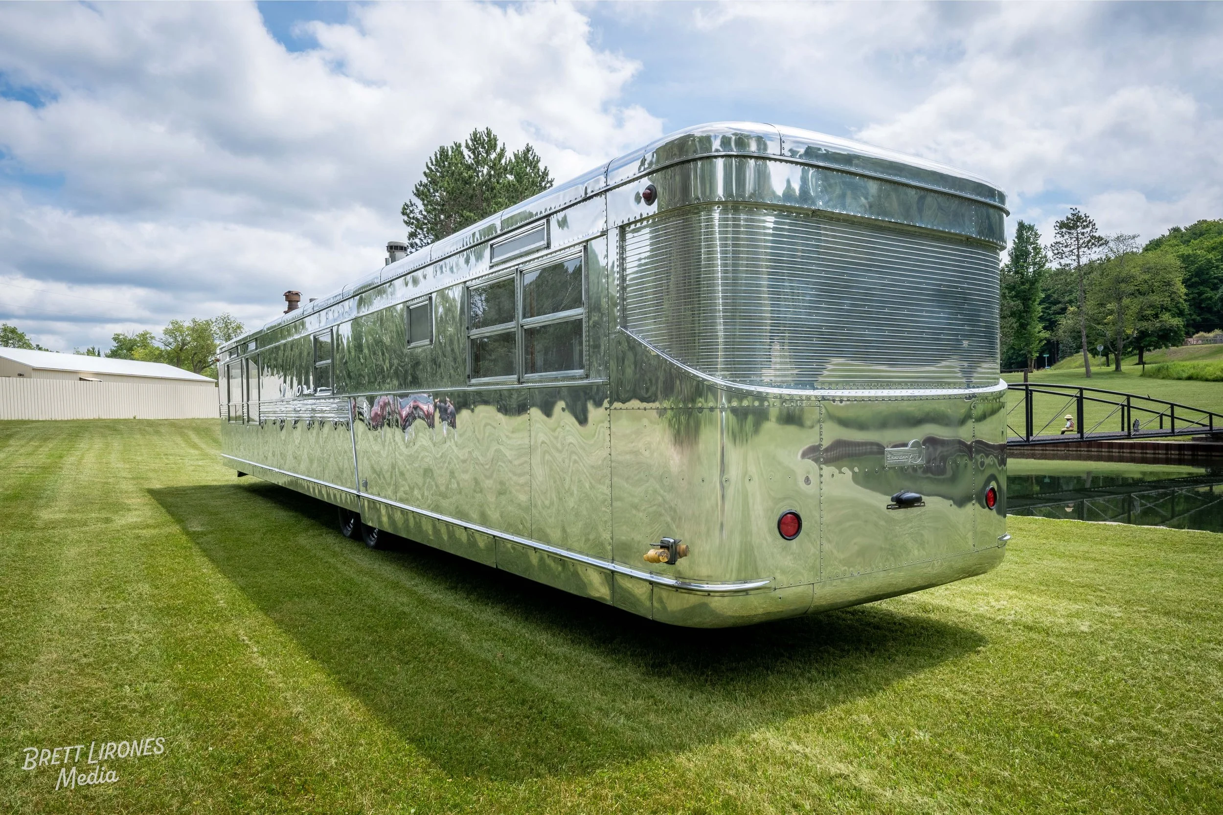 A shiny, metallic food truck parked on a grassy area with trees and a small body of water in the background.
