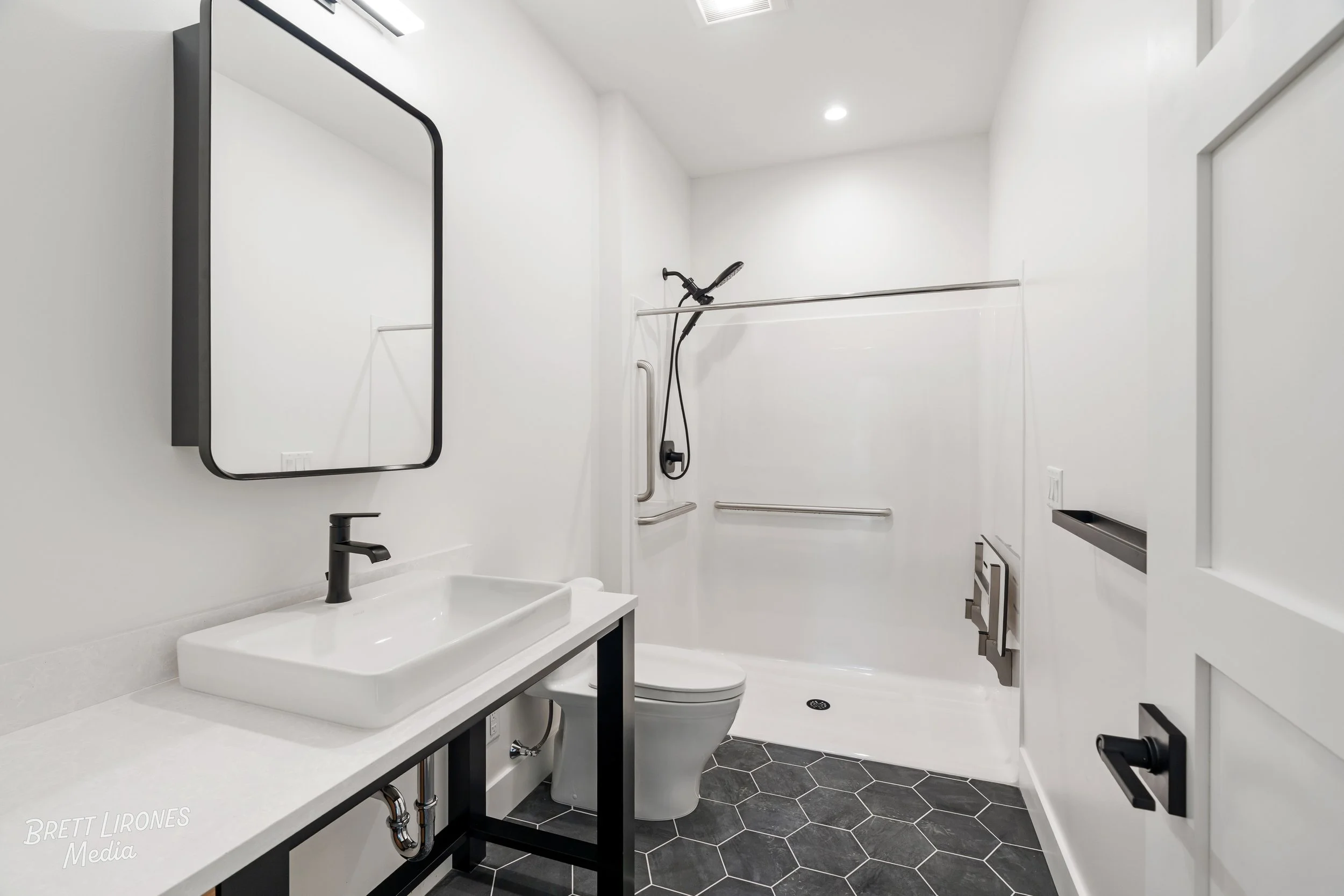 Modern bathroom with a white sink on a black stand, a large mirror, a toilet, and a walk-in shower with black fixtures and grab bars, black hexagonal floor tiles, and white walls.