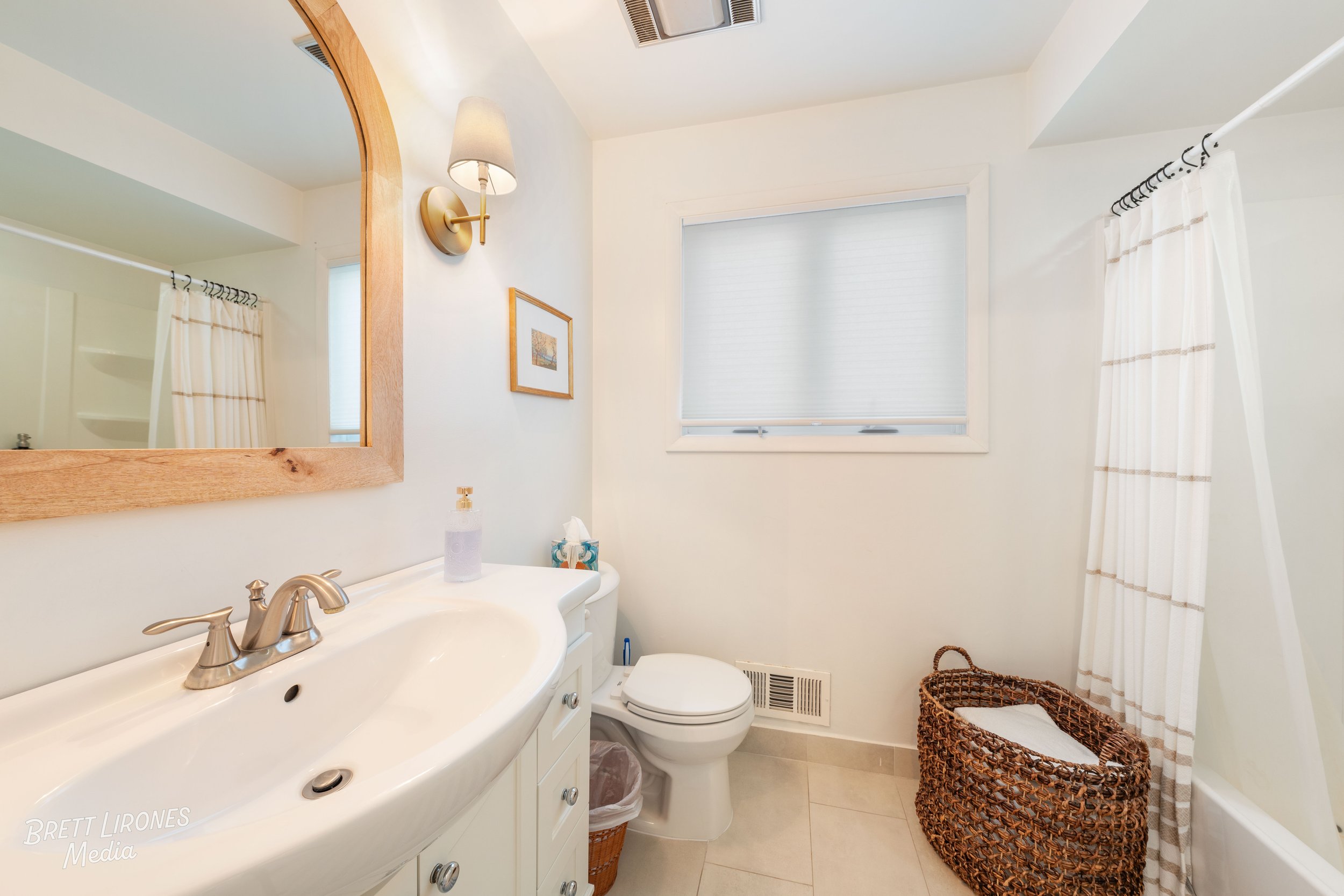 A small bathroom with a white sink, wooden-framed mirror, a toilet, a window with blinds, a woven laundry basket, a shower curtain, and wall-mounted light fixtures.