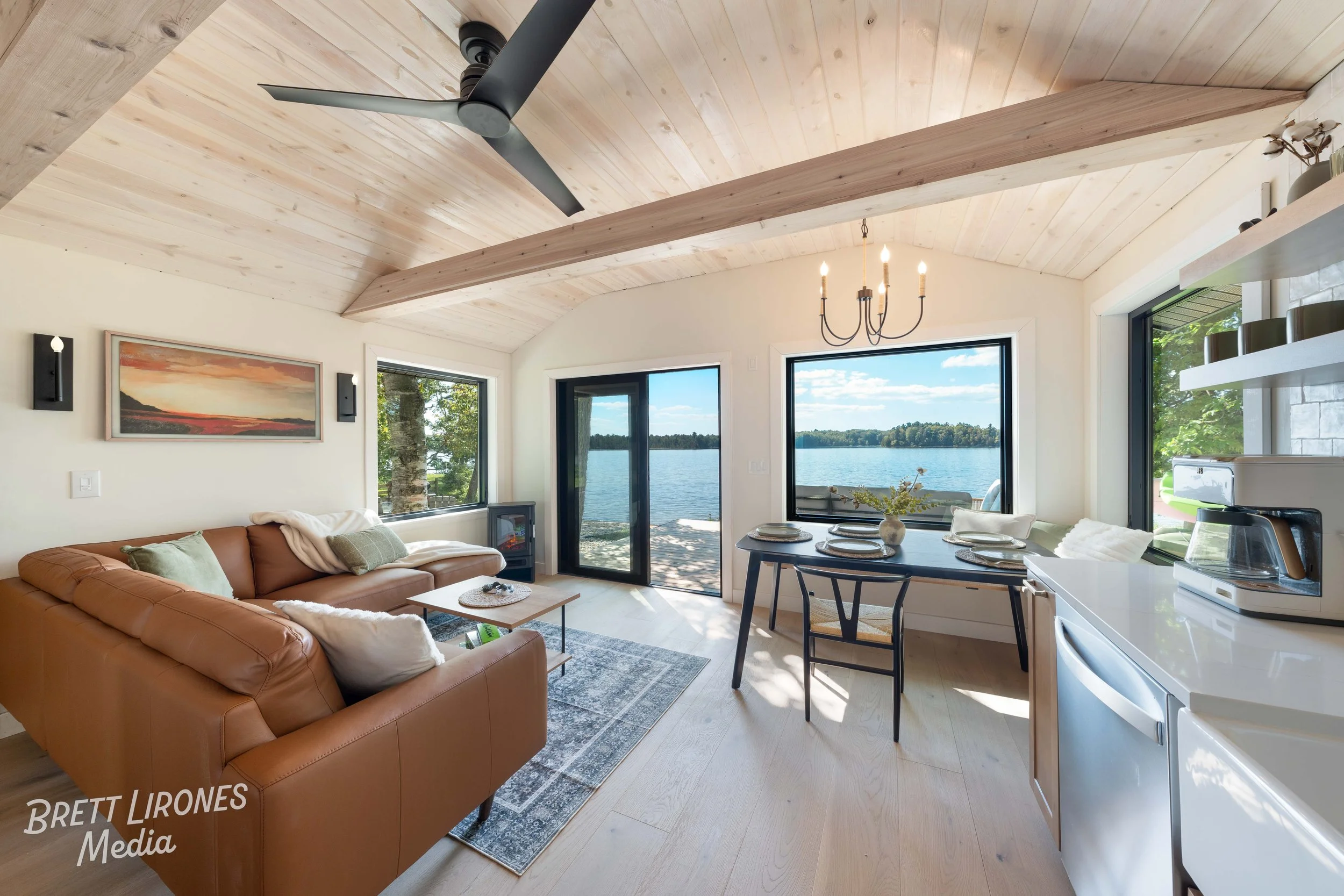 Living room with large windows and a view of a lake, featuring a brown leather sofa, wooden coffee table, a rug, a dining table with plates, and a kitchenette, with a ceiling fan and chandelier.