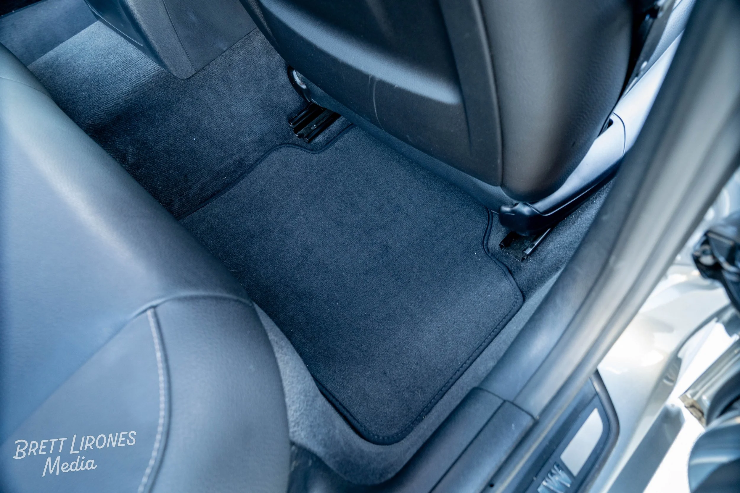 The back seat area of a vehicle showing black carpeted floor mats and portions of black leather seats.