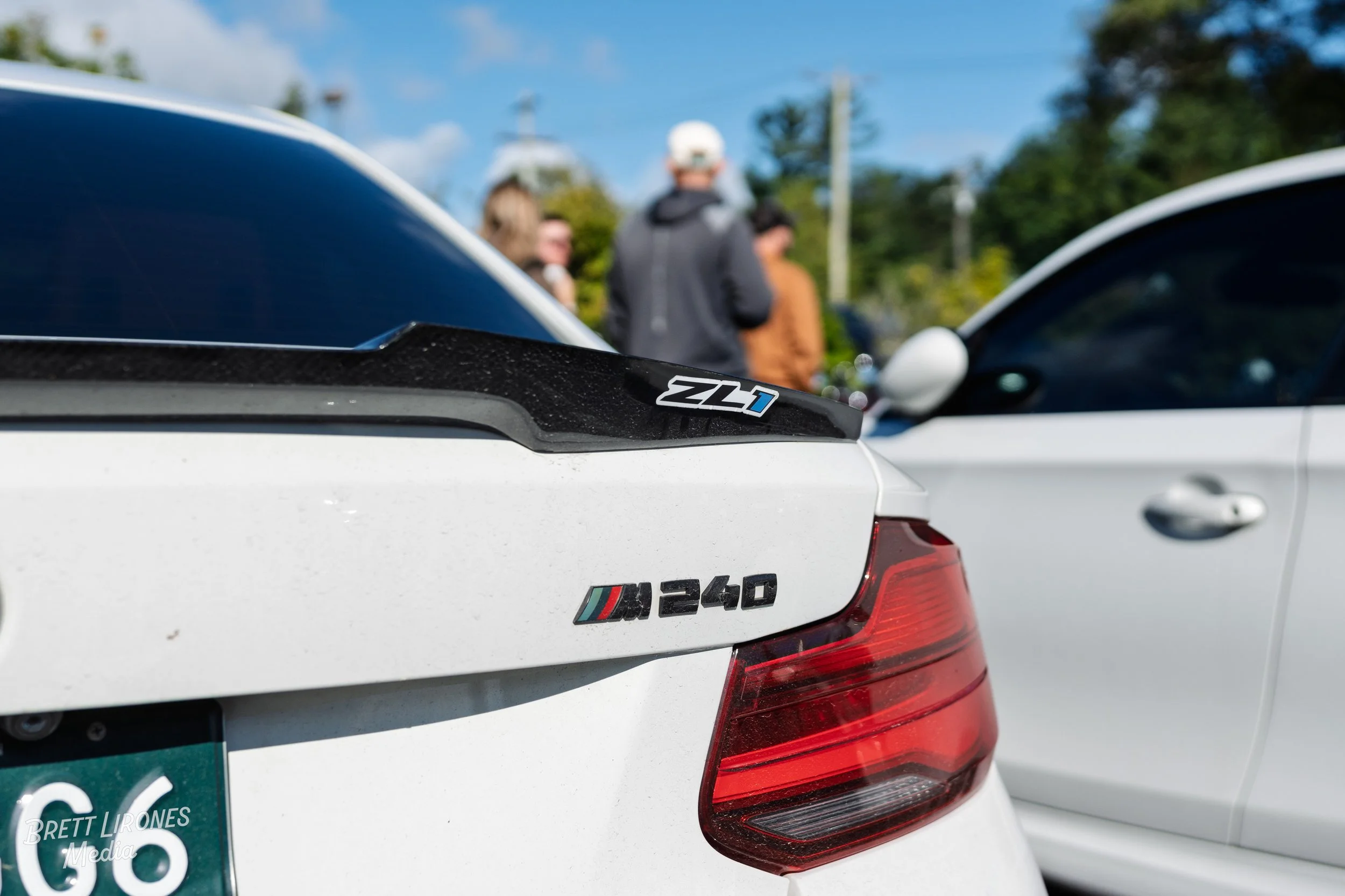 Close-up of a white BMW M240 with an ZL1 badge on a black spoiler, parked outdoors. People are blurred in the background, and part of another white car is visible to the right.