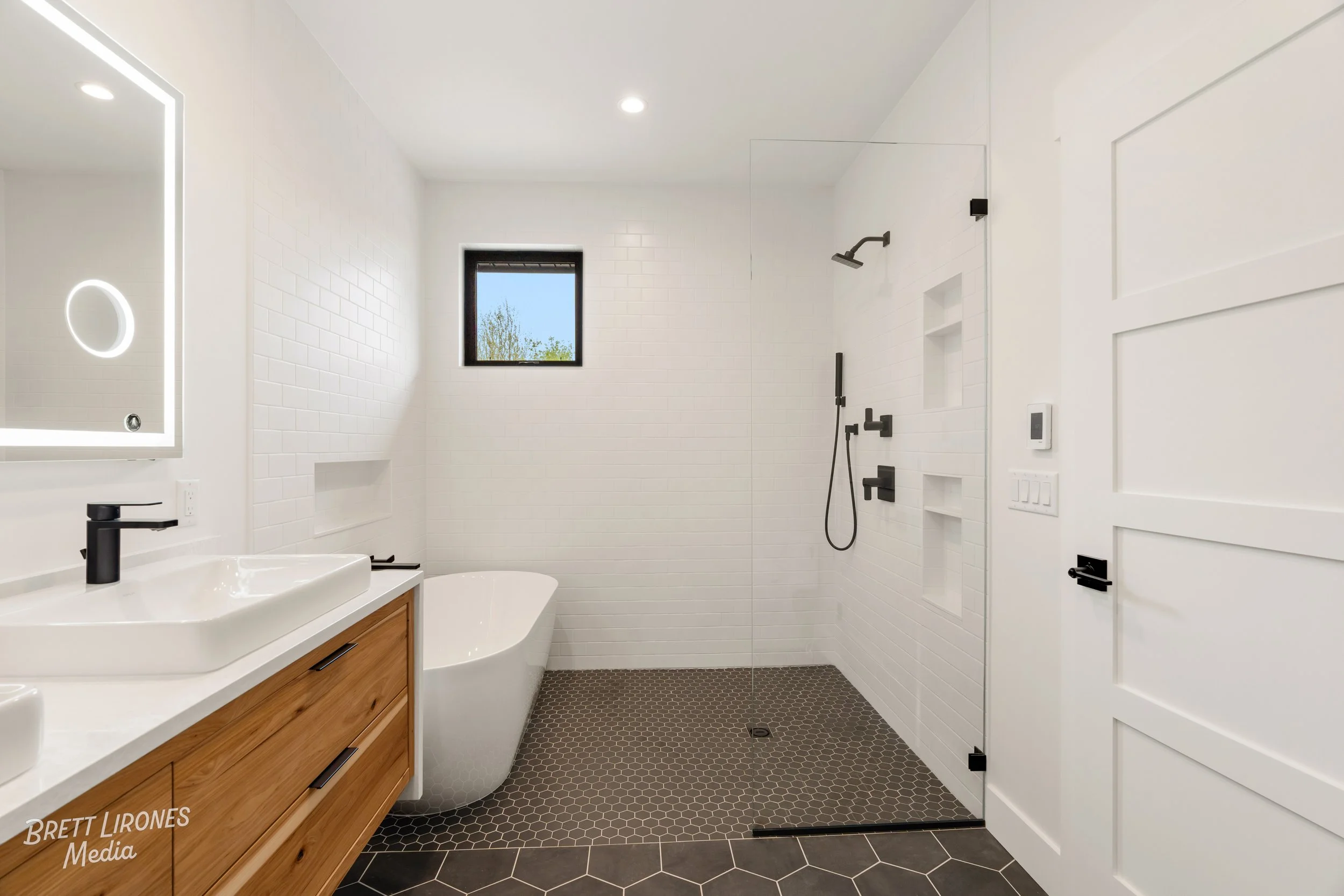 Modern bathroom with white walls, black fixtures, a wooden vanity, a freestanding bathtub, a walk-in shower with black fixtures, a small window, and a gray tile floor.