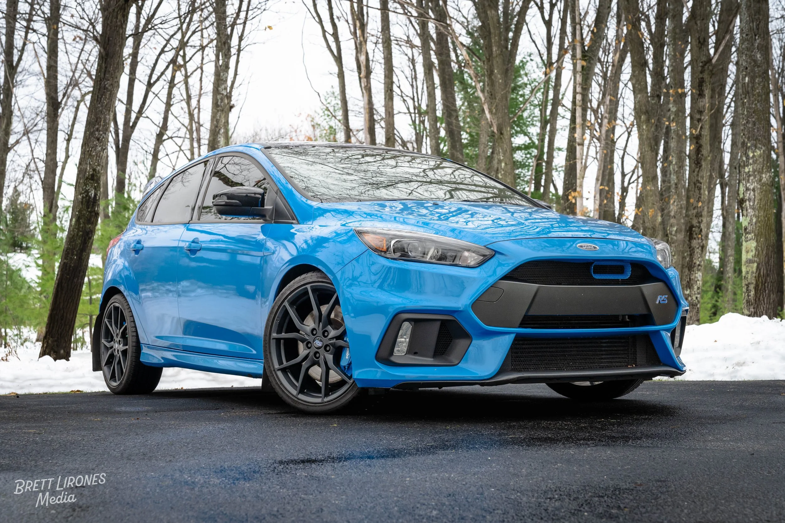 A blue Ford Focus RS parked on a paved surface in a wooded area with some snow on the ground.