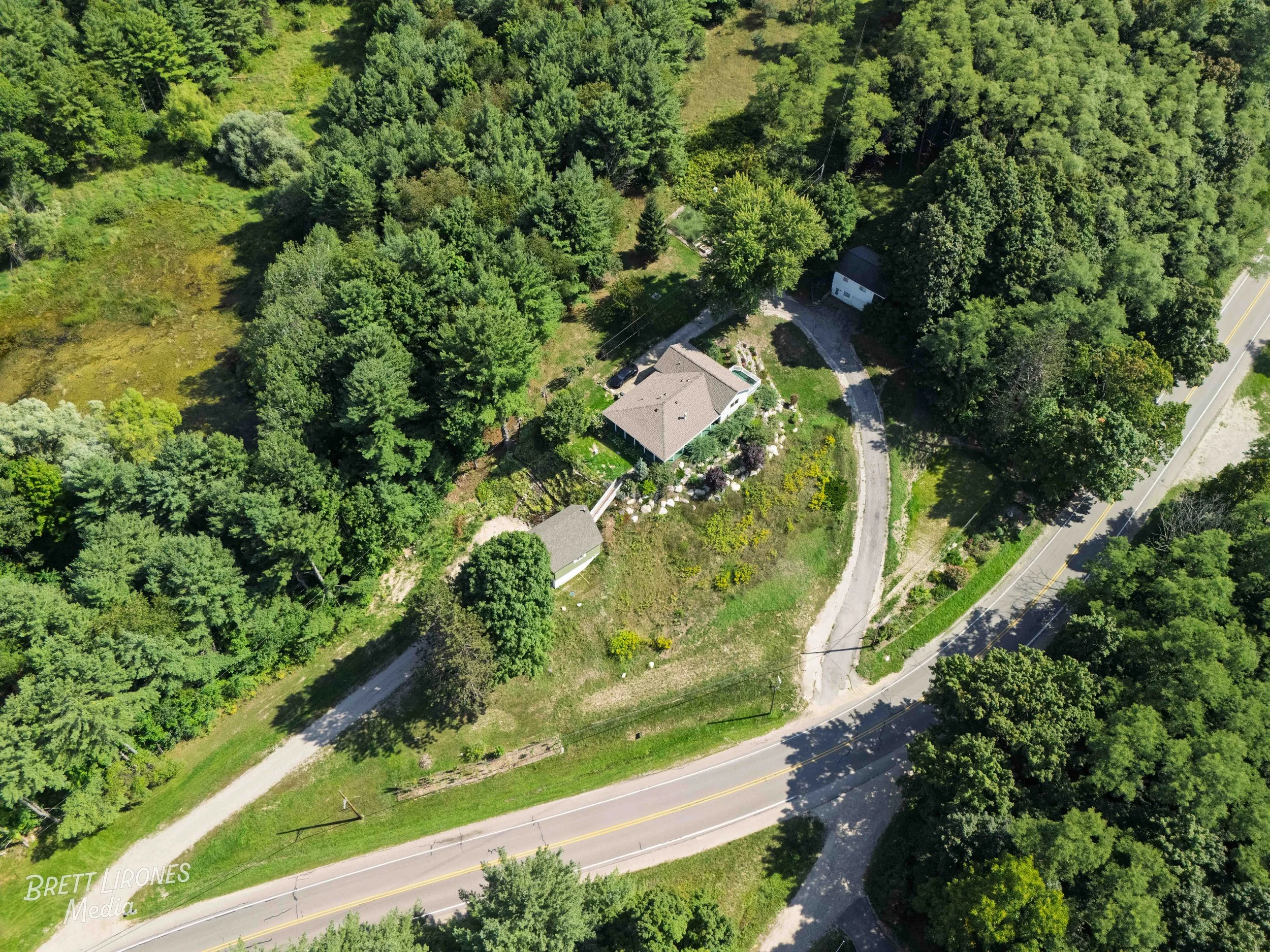 Aerial view of a house surrounded by trees and greenery, with a driveway, a small shed, and a road nearby.